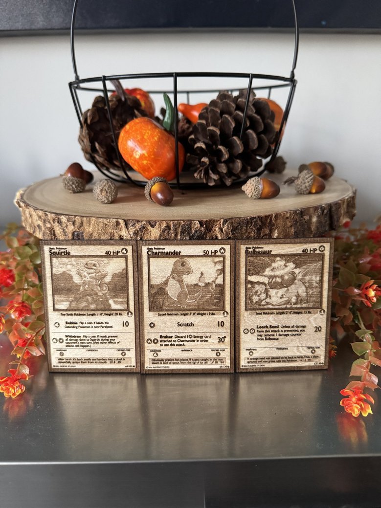 A decorative display featuring a wooden slab topped with a black wire basket filled with pinecones and small decorative objects. In front, three vintage Pokémon cards are propped up on the slab, with artificial red and orange flowers around the edges