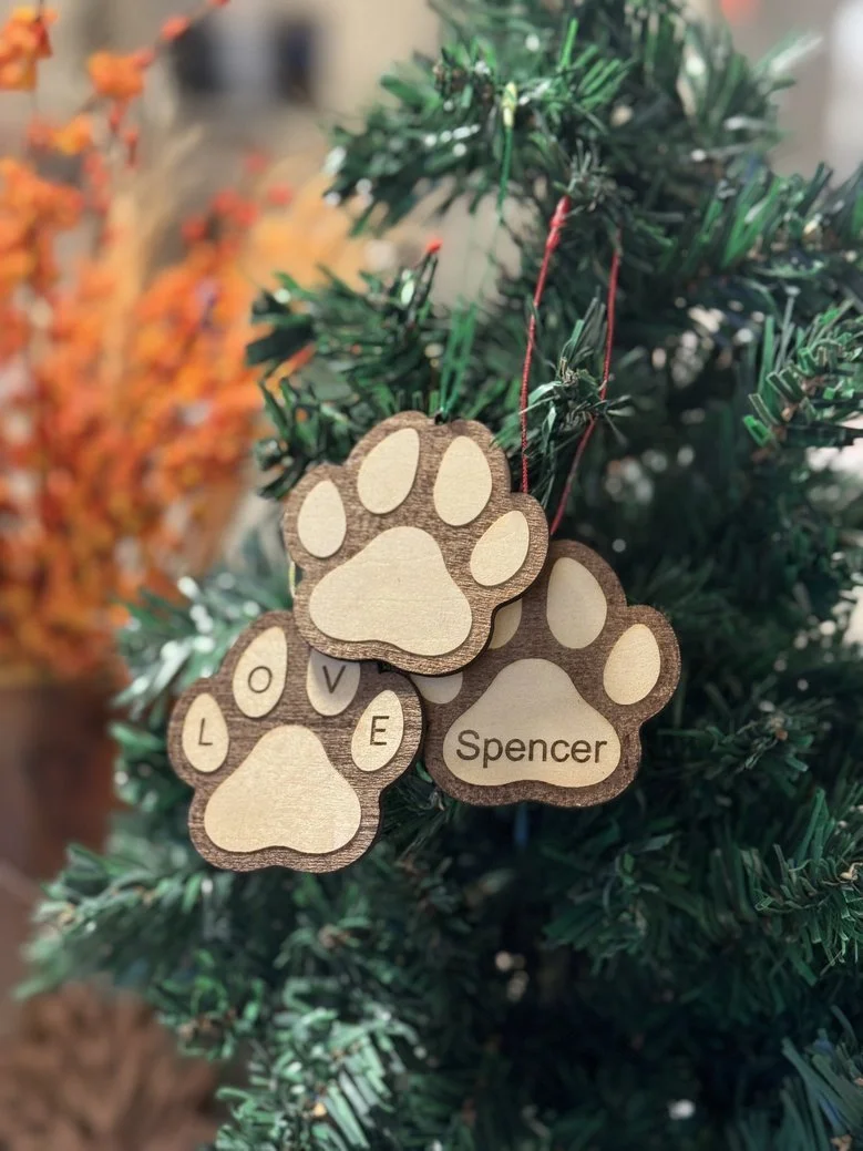 Wooden Christmas tree ornament shaped like a paw print with the name 'Spencer'. It is hanging on a Christmas tree branch with orange and red decorations visible in the background.