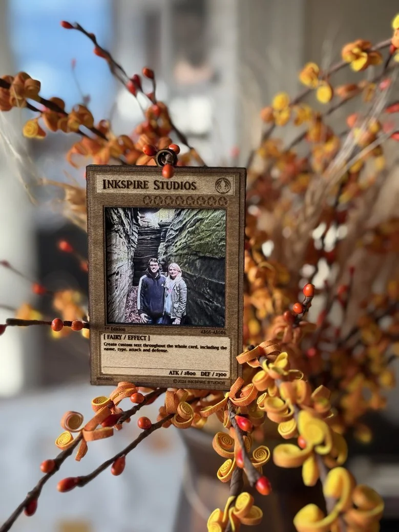 A custom-designed fantasy card from Inkspire Studios hanging from a branch of orange and yellow decorative curly twigs with red berries, with a blurred background.