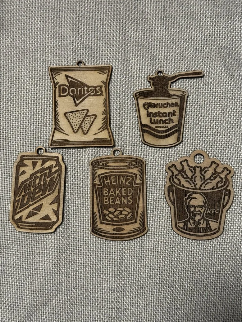 Five wooden keychains with logos of popular fast food and beverage brands, including Doritos, Maruchan Instant Lunch, Mountain Dew, Heinz Baked Beans, and KFC.