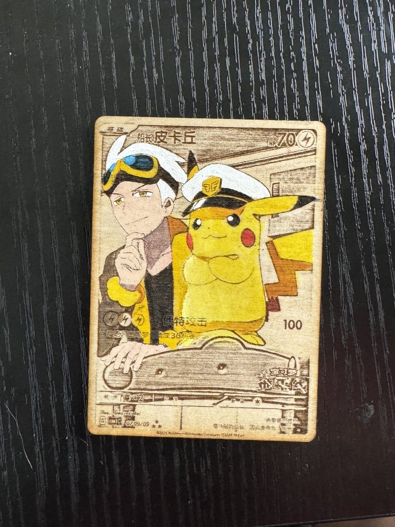 A wooden card featuring a printed illustration of a young man with goggles and a Pokémon character Pikachu dressed as a pilot. The card has various Japanese characters and symbols, along with some numbers, set against a black textured surface.
