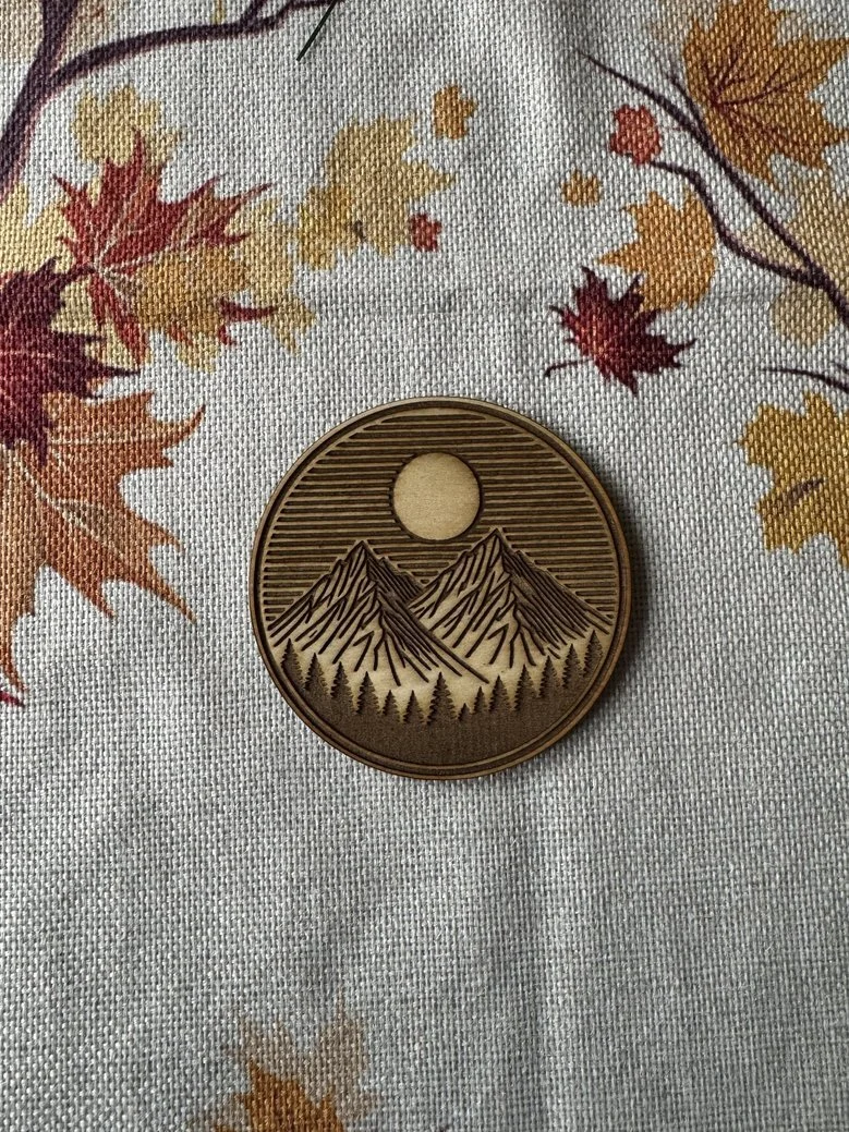 Round wooden medallion with engraved mountains, trees, and a sun or moon, placed on autumn leaf-patterned fabric.