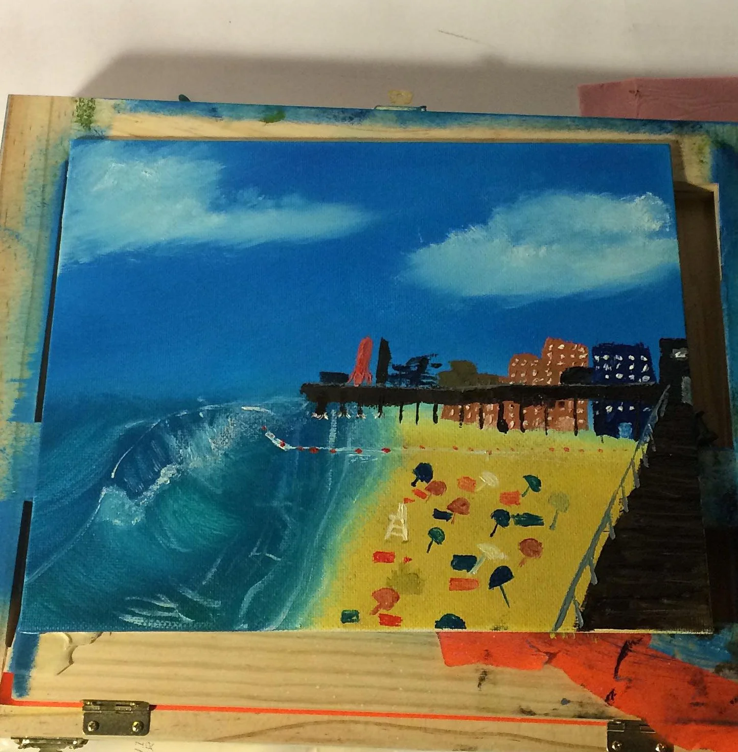 Painting of a beach scene with blue sky, clouds, buildings, pier, and colorful beach umbrellas and chairs.