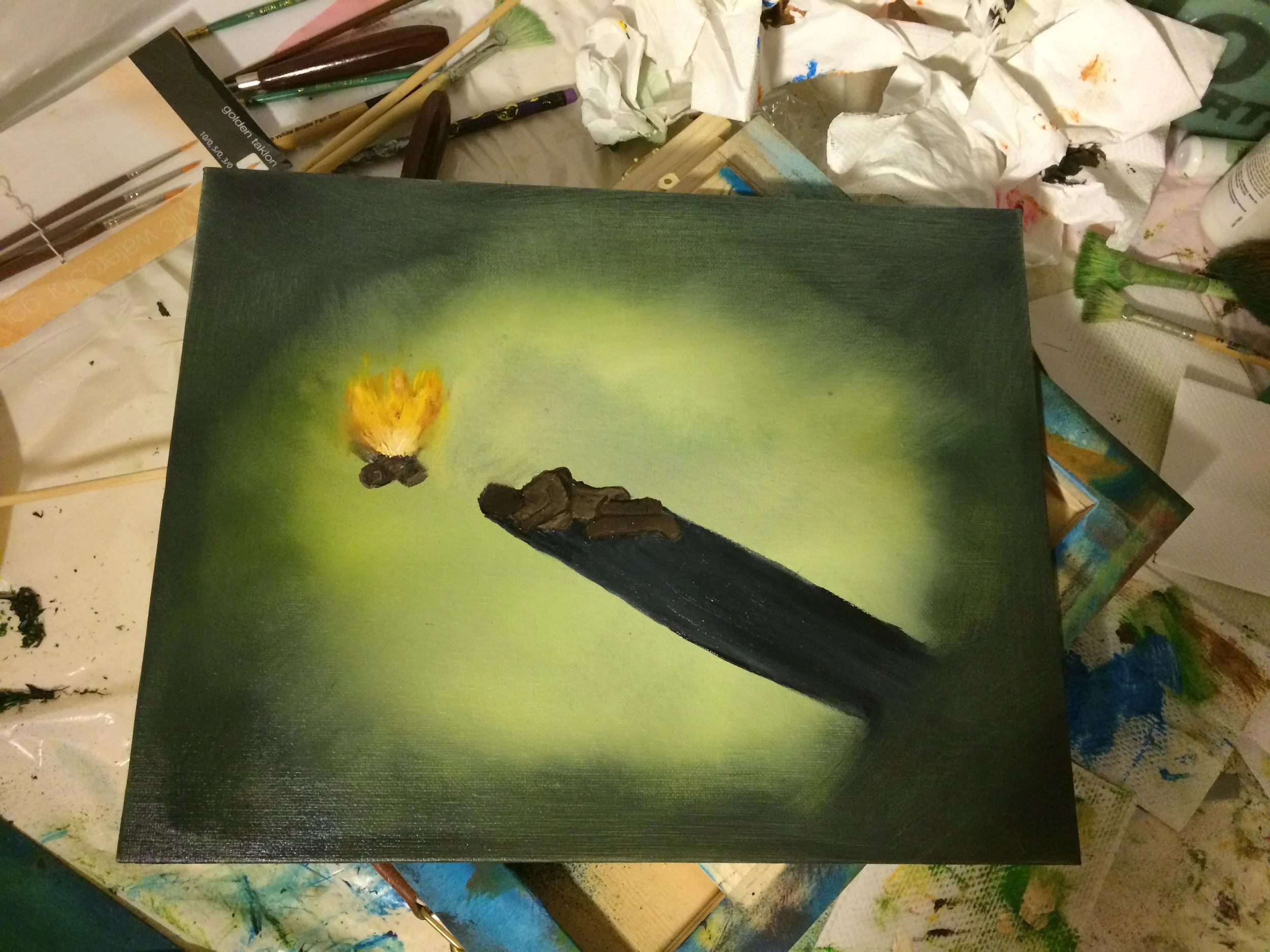 A small painted canvas depicts a campfire with flames and a short wooden log against a green gradient background, surrounded by painting supplies and cluttered workspace.