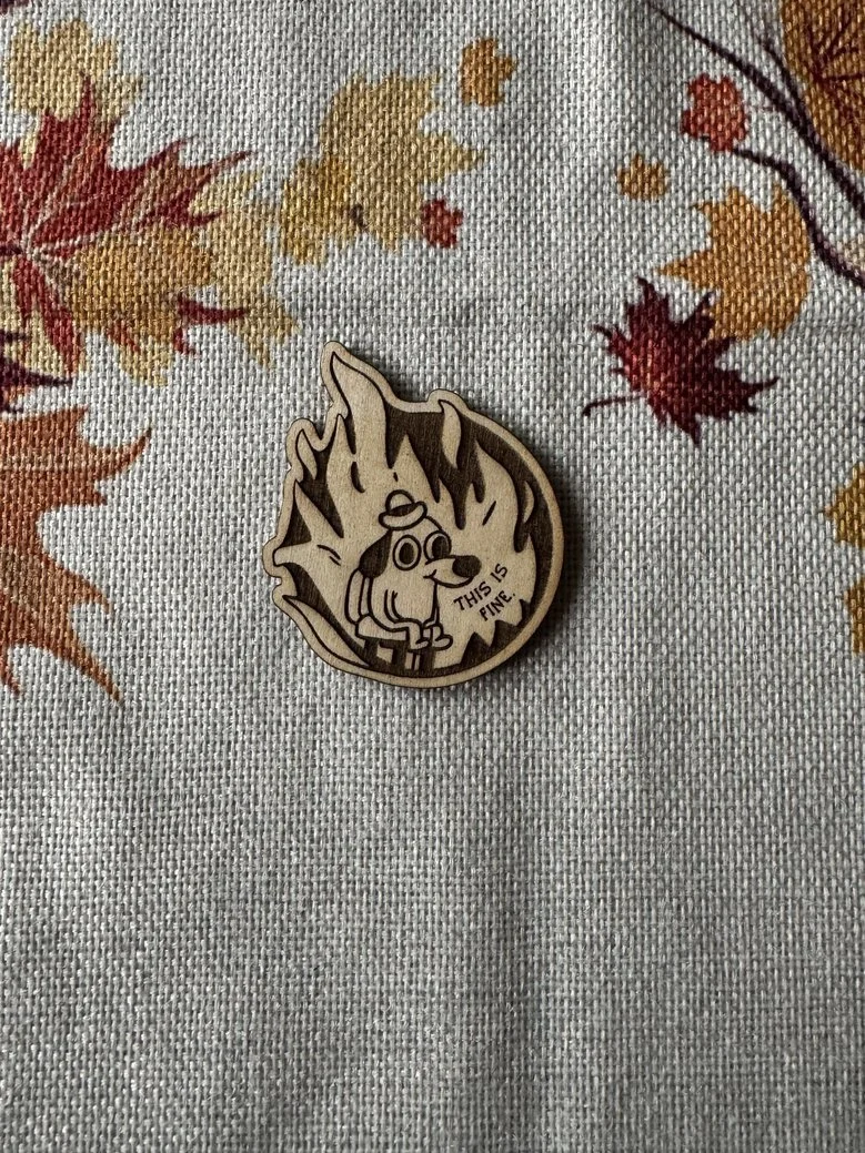 A wooden pin featuring a cartoon dog with a fire behind it. The dog has a worried expression and a speech bubble that says "This is fine". The pin is placed on a fabric with a colorful autumn leaf pattern.