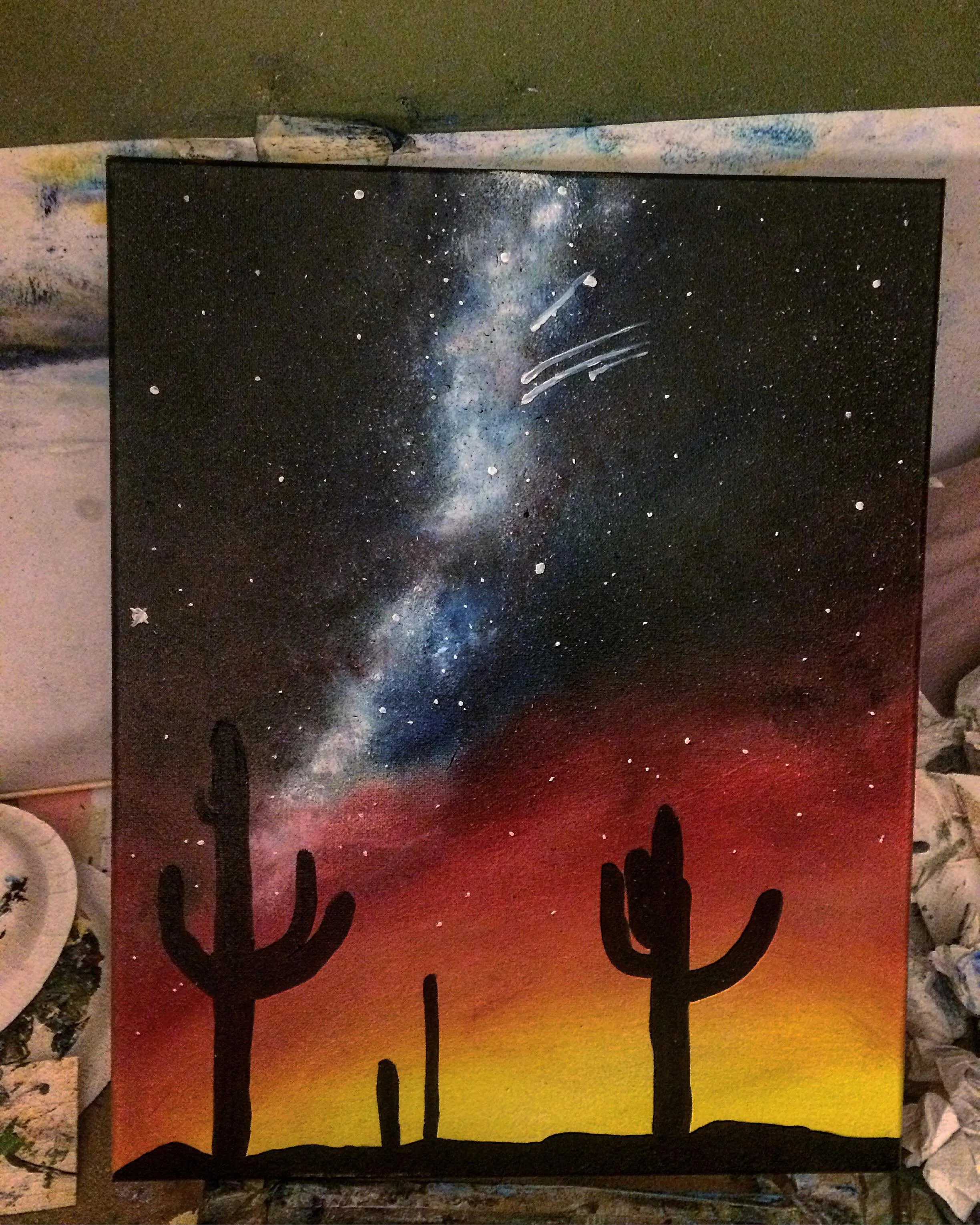 Painting of a desert landscape at sunset with silhouettes of cacti, a colorful sky transitioning from yellow to red, and a starry night sky with the Milky Way galaxy and shooting stars.