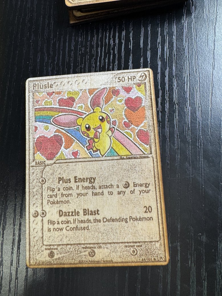 Pokemon card featuring Pikachu with a colorful background of hearts and rainbows. The card shows Pikachu with large eyes, pink cheeks, and a cheerful expression, surrounded by hearts. It is titled 'Plusle' and has two attacks: 'Plus Energy' and 'Dazz