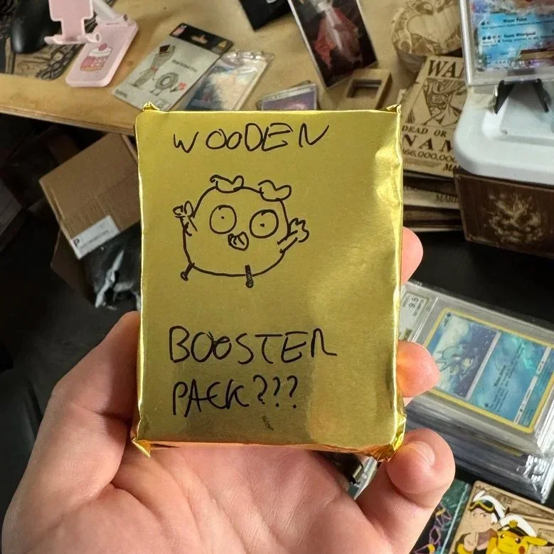 Hand holding a gold-wrapped package with handwritten notes and a drawing of a pig on it, surrounded by other trading cards and items on a table.