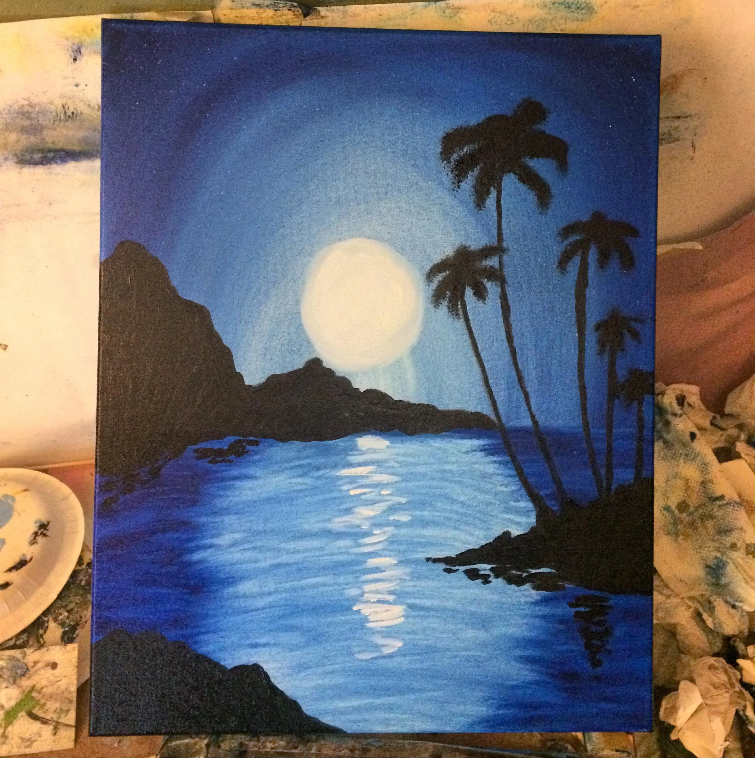 A painting of a moonlit coastal scene with a large full moon in the sky, reflecting on calm water, with silhouetted palm trees and rocky formations. The painting uses shades of blue, black, and white.