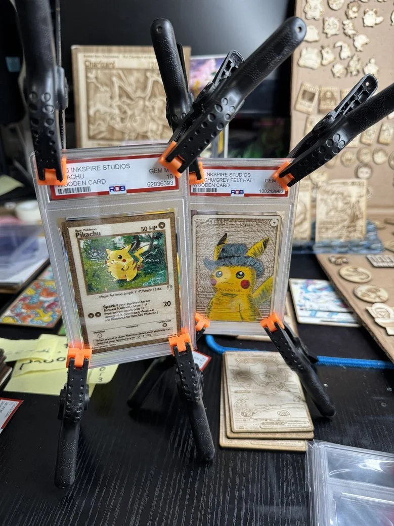 Two graded Pokémon cards, a Pikachu card and a Pikachu illustration, secured with black clips on a display stand, with various other card packs and collectibles in the background.