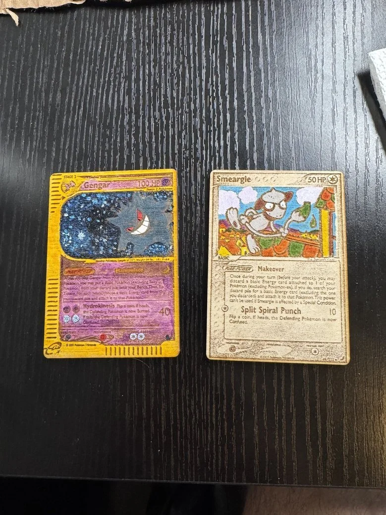 Two Pokémon trading cards, Gengar on the left and Smeargle on the right, laid flat on a black wooden surface.