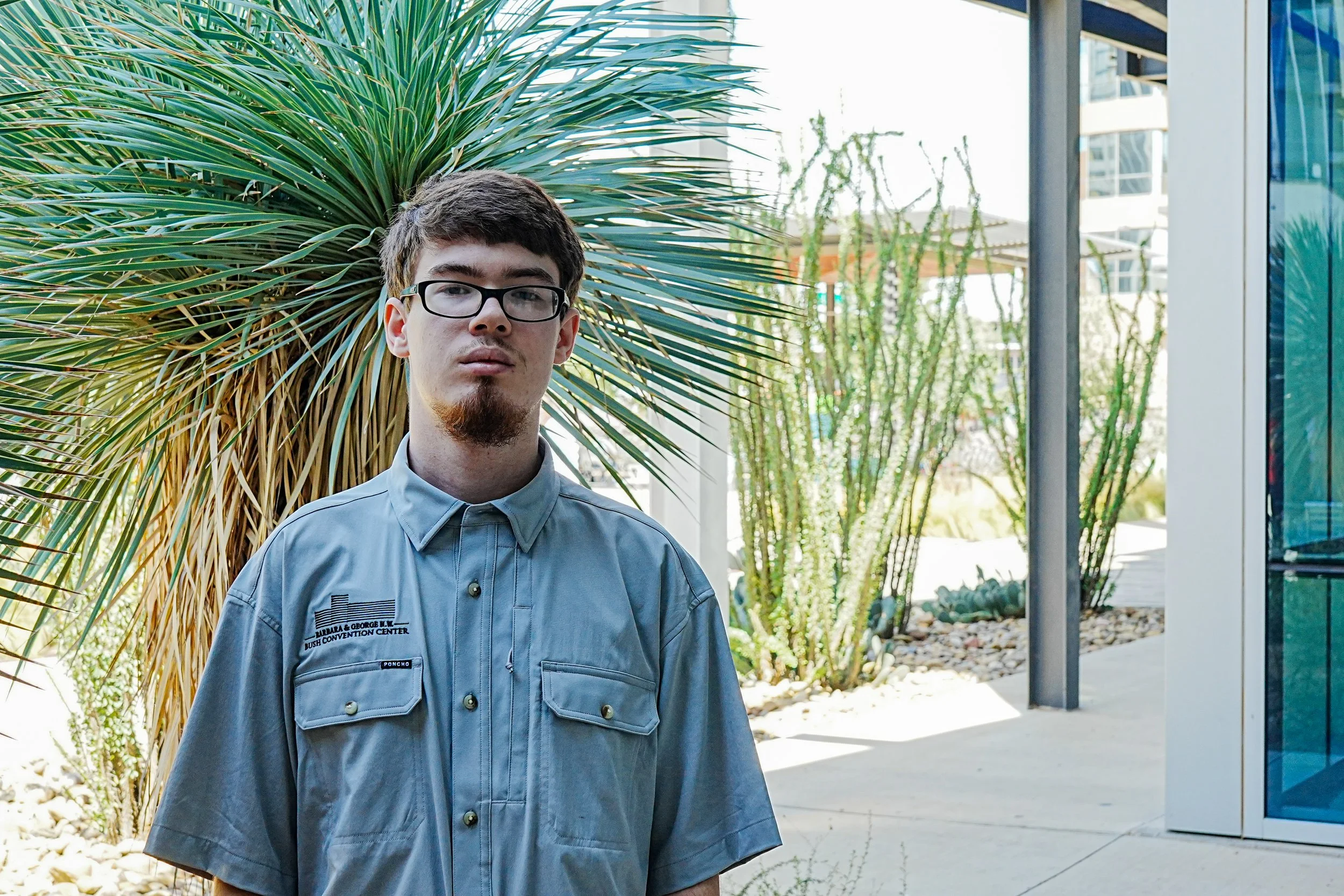Young man with glasses, light skin, brown hair, and facial hair wearing a grey work shirt with patches, standing outside in front of a glass building with trees in the background.