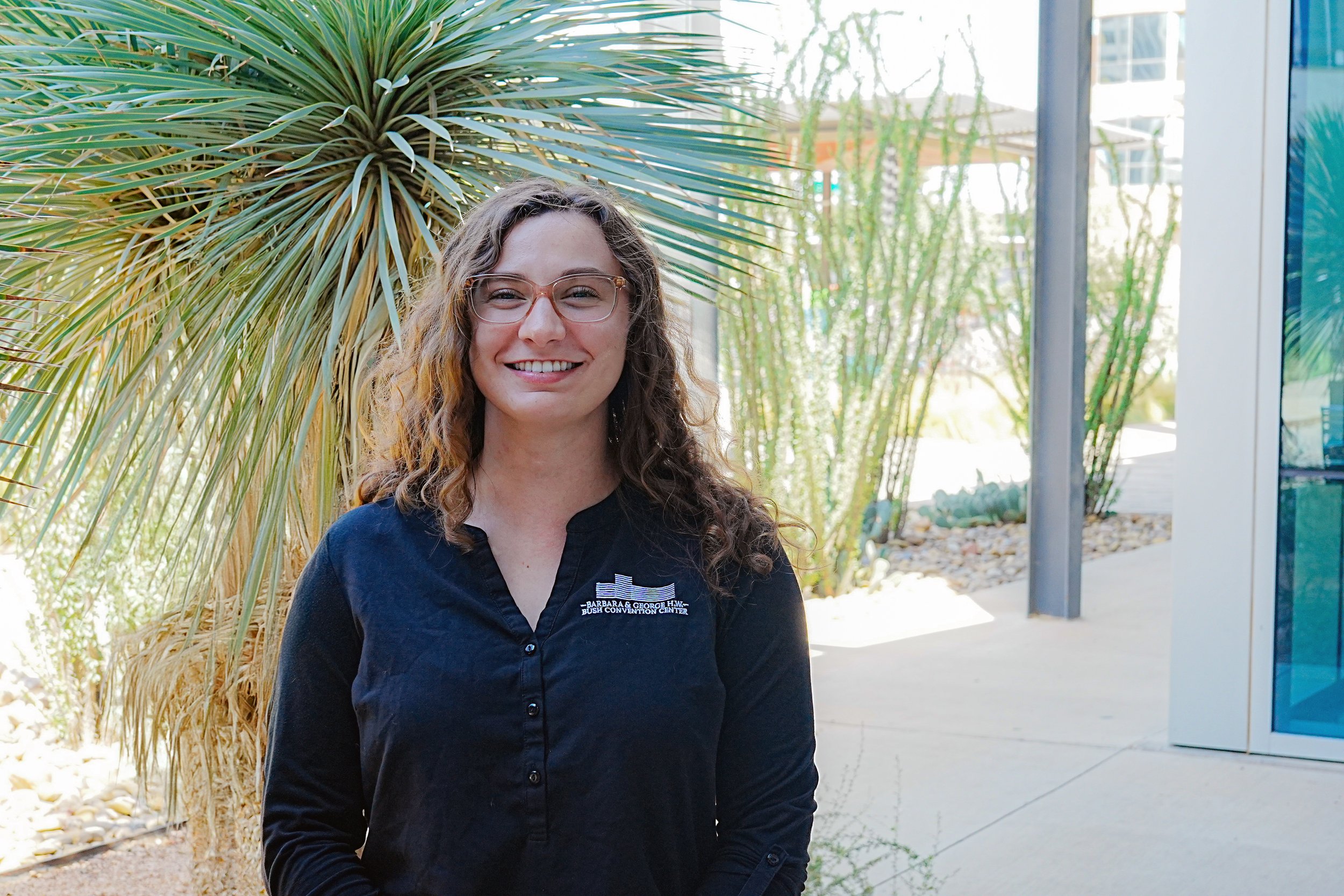 A woman with curly brown hair and glasses, wearing a navy uniform, smiling outdoors in front of a glass building.