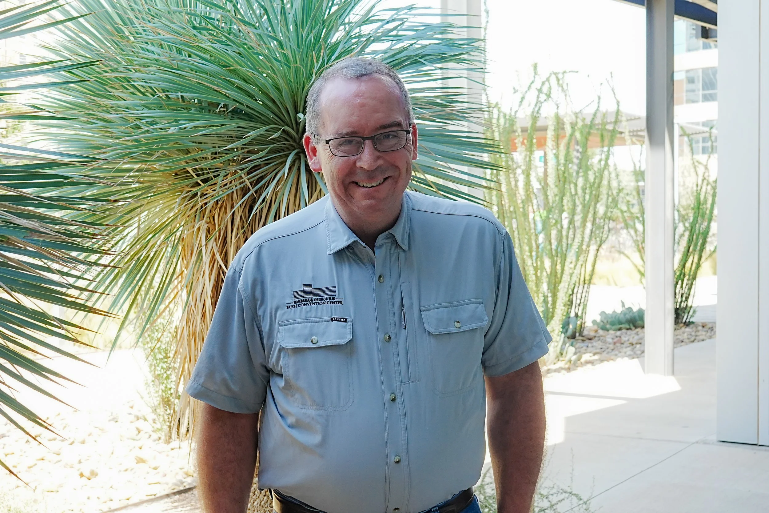A man with glasses smiling outdoors in front of modern buildings, wearing a button-up shirt.