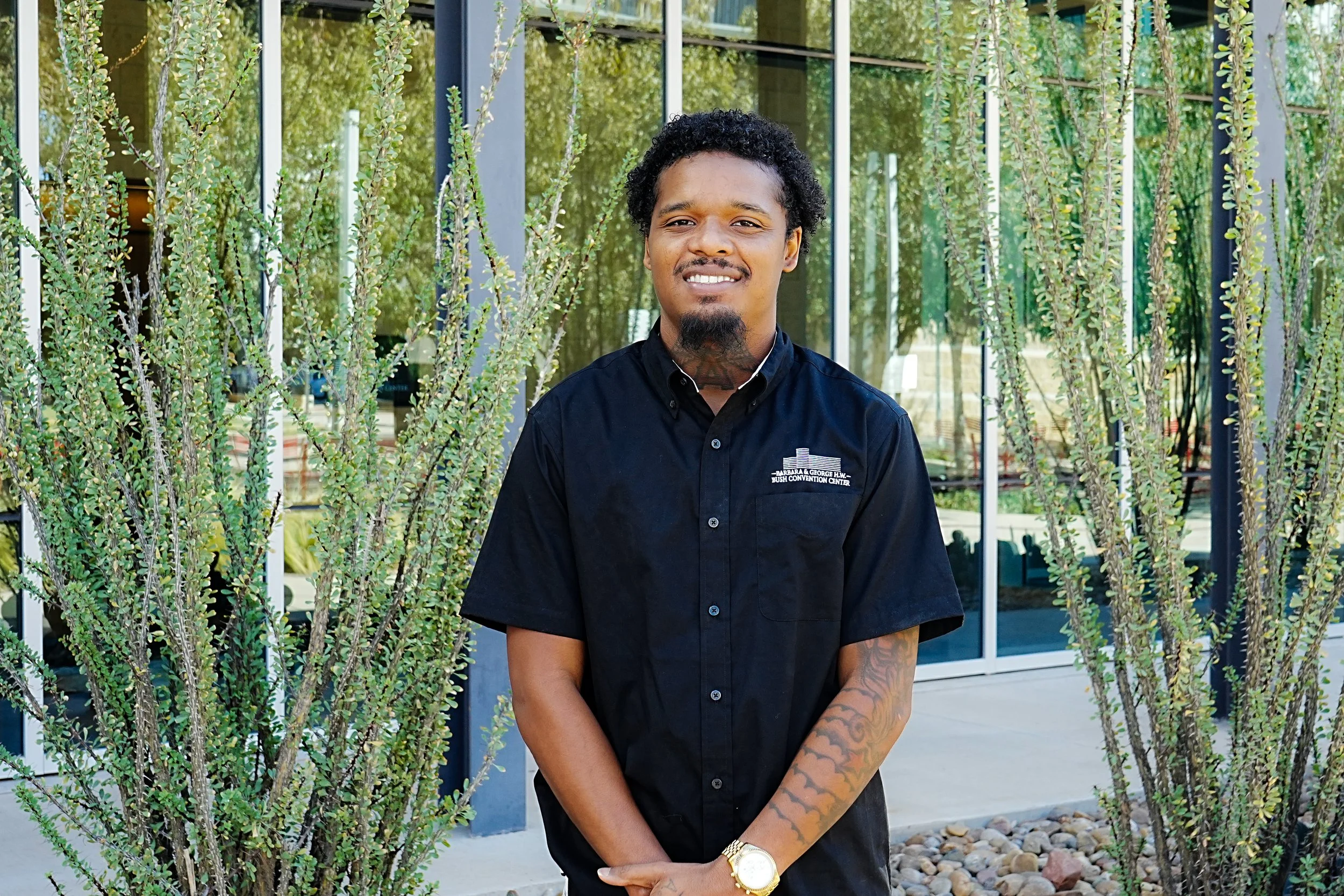 A man standing outdoors in front of a glass building with tall desert plants, wearing a black button-up shirt with logo, a gold watch, and smiling.
