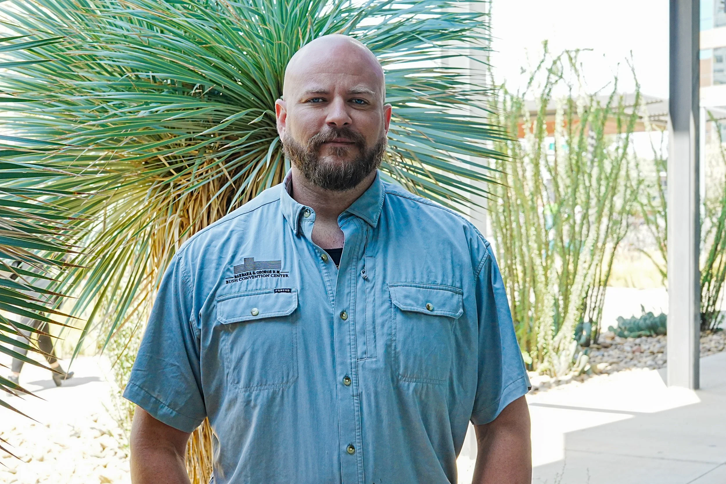 A man with a beard and shaved head smiling outdoors in front of a building with tall plants.
