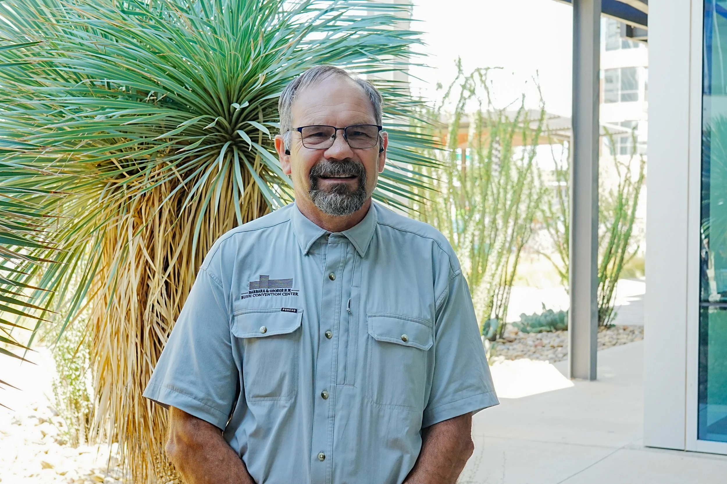 A man with glasses, a beard, and a mustache smiling outdoors in front of a modern building.