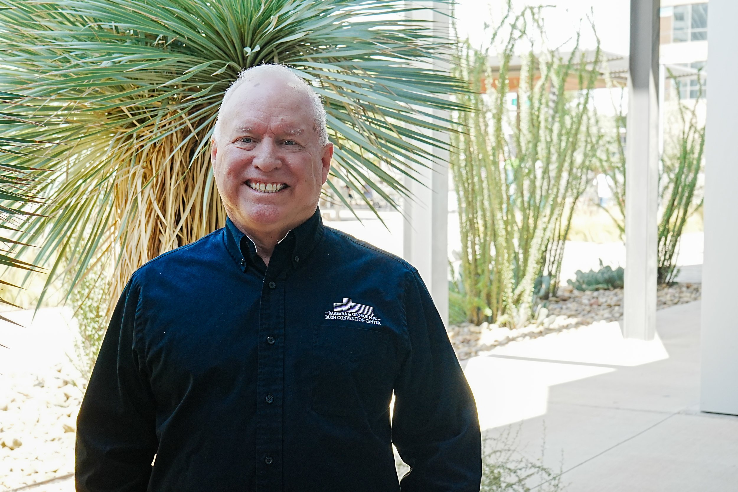 A man with short blond hair wearing a blue collared shirt and dark blue vest, smiling outdoors with a wooden fence in the background.