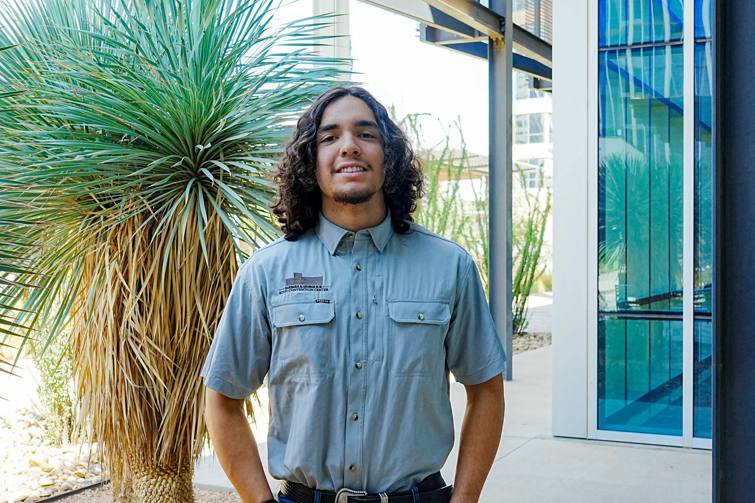 A young man with long, curly hair and a goatee, wearing a gray collared shirt with a logo, standing outside near a glass building and trees.