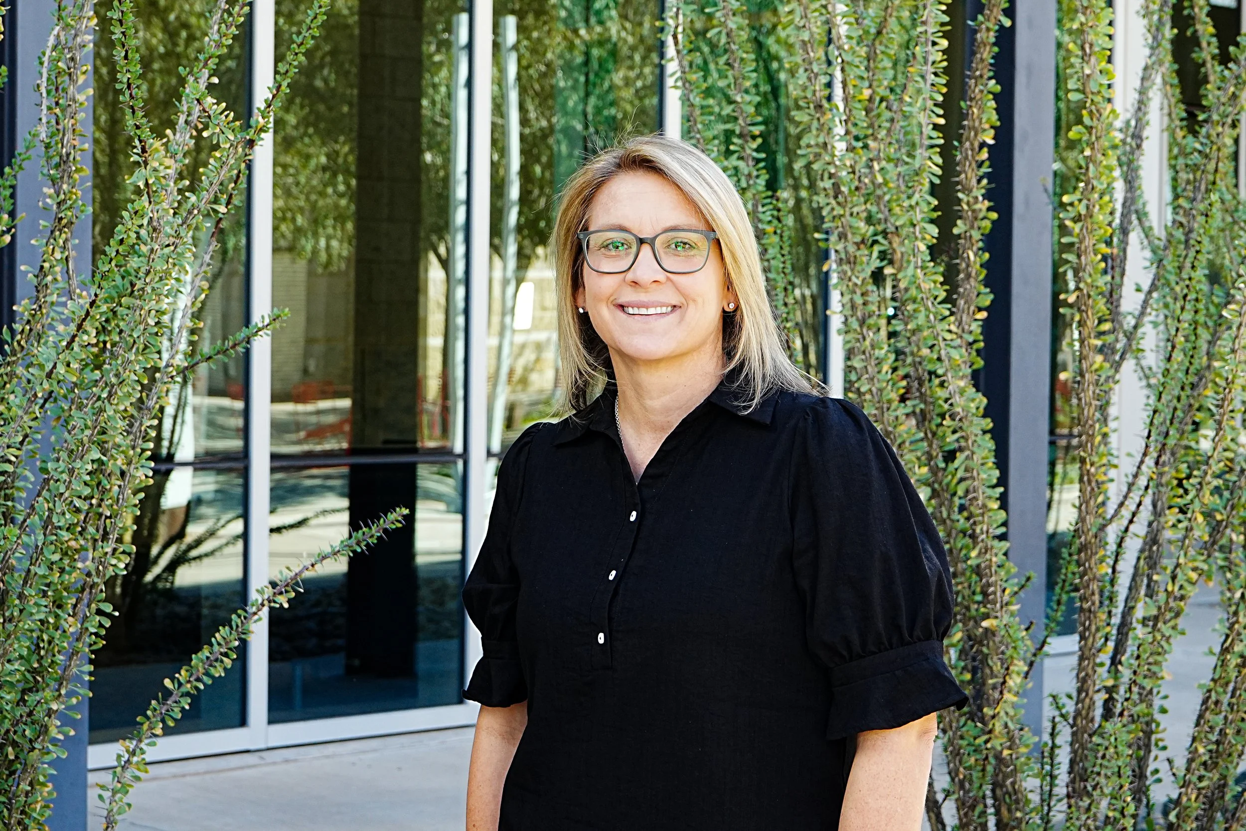 A woman with blonde hair and glasses smiling, standing outdoors near a glass building.