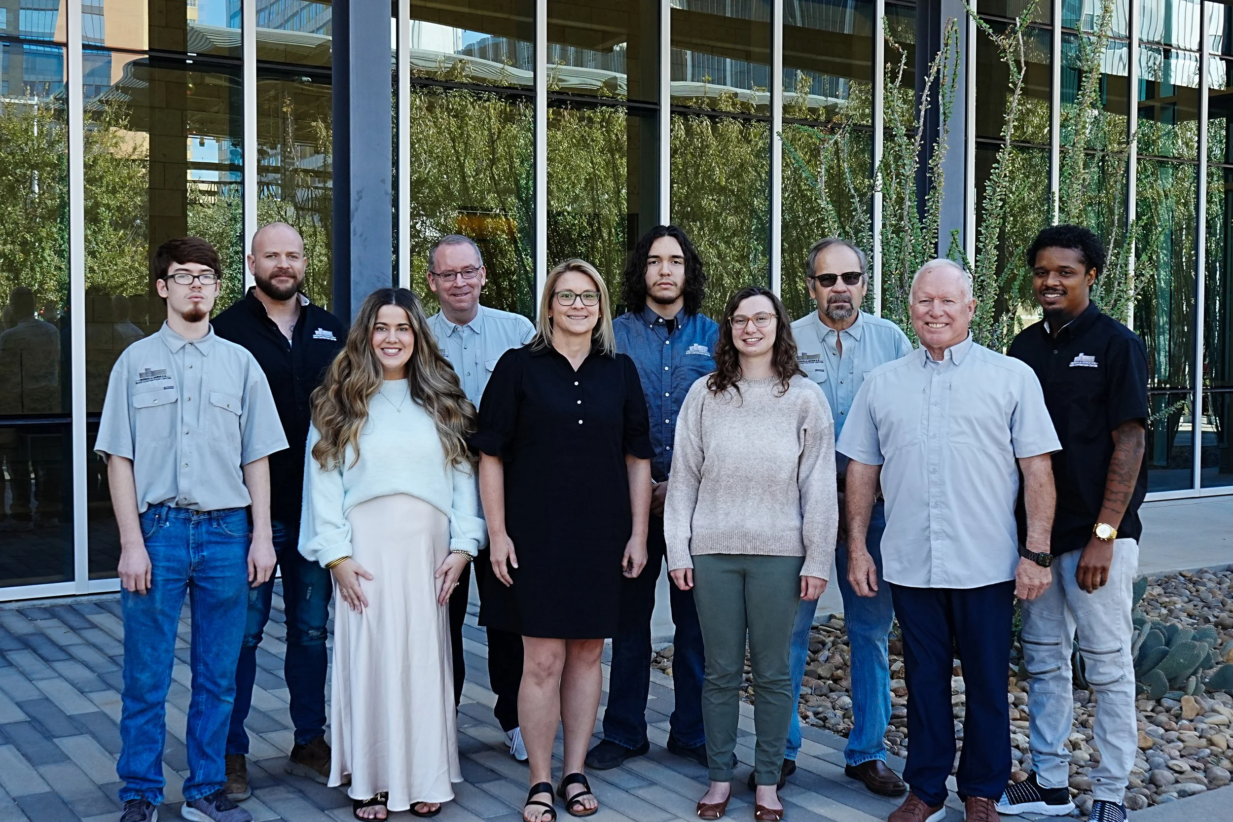 Group photo of eleven people standing outside in front of a glass building, with some trees and desert plants visible in the background.