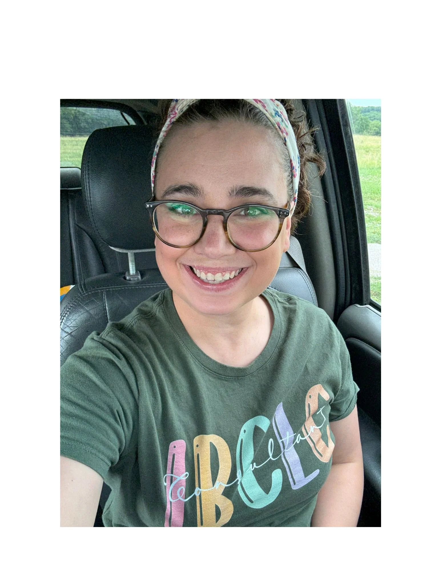 A smiling woman with curly brown hair, glasses, and a colorful headband, wearing a green T-shirt with the word IBCLC in colorful letters, sitting inside a vehicle with a countryside background visible through the window.