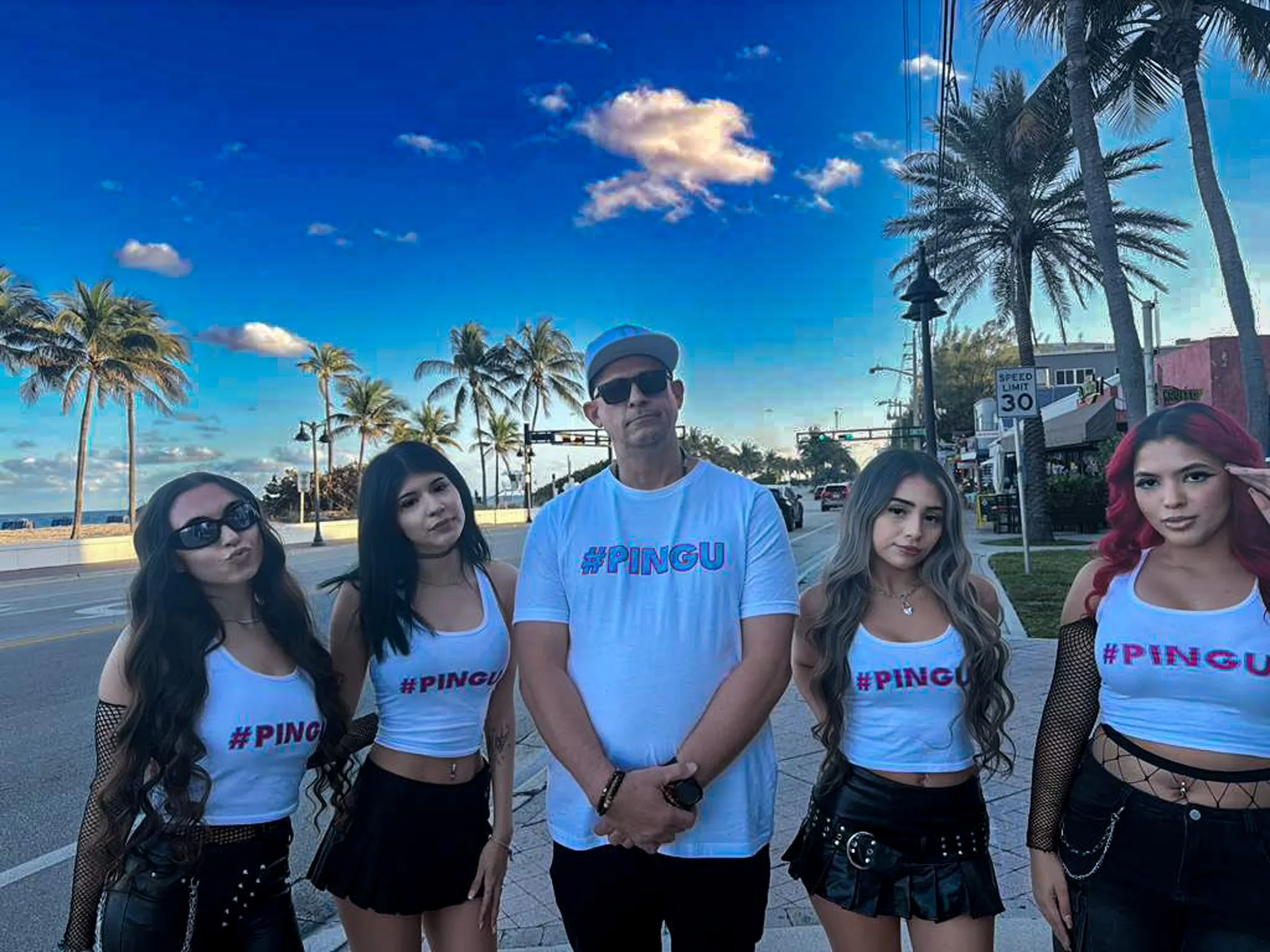 Group of four women and one man standing on a city sidewalk with palm trees and a blue sky in the background. All are wearing white T-shirts with '#PINGU' printed in pink and blue.