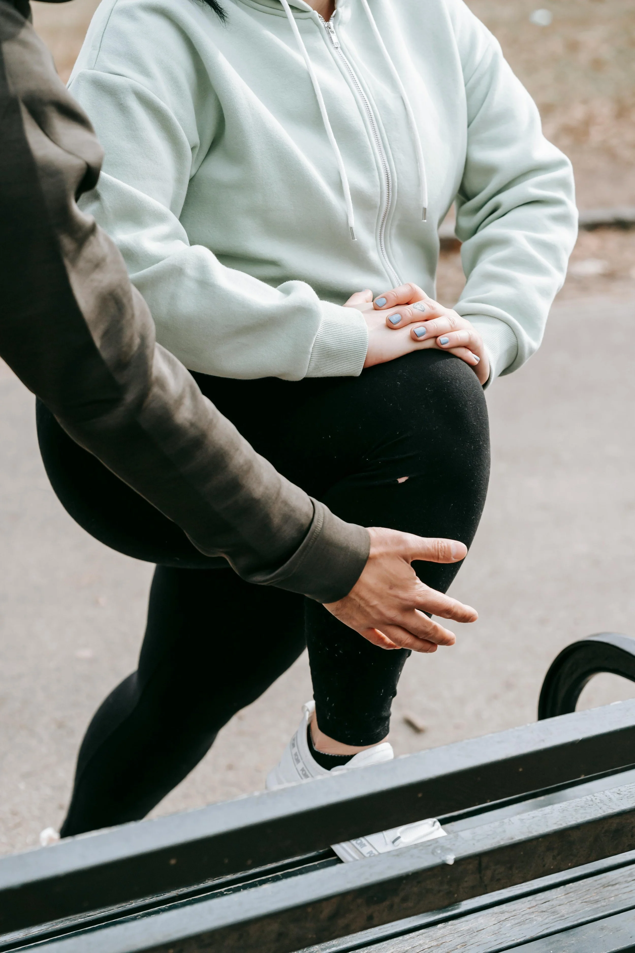 A person in a gray hoodie and black leggings is assisting another person with a knee injury, holding their leg on a park bench outdoors.