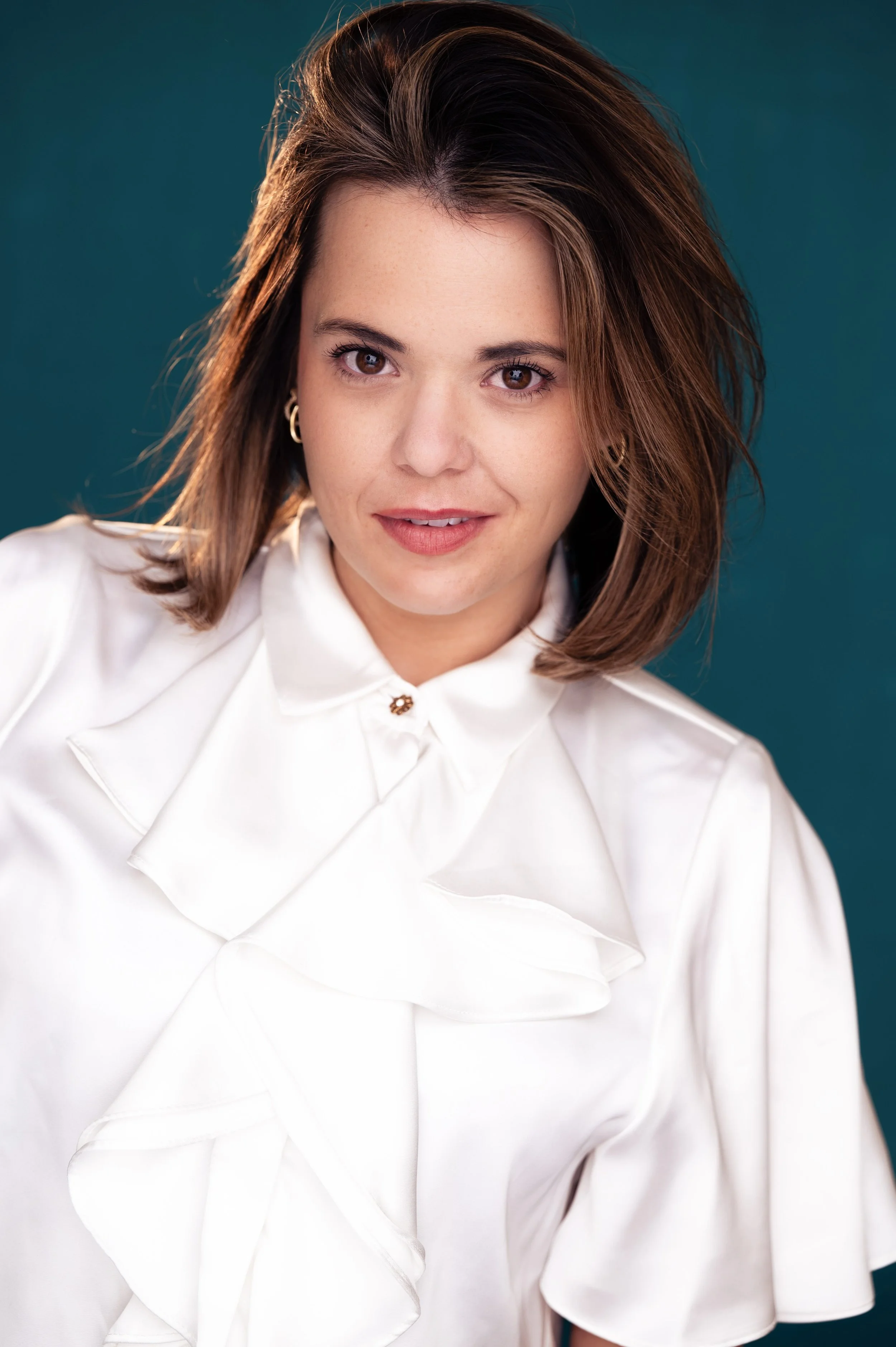 A woman with shoulder-length brown hair and fair skin, sitting on a wooden stool against a plain brown backdrop, wearing a sleeveless denim top with lace-up front and wide-legged maroon pants.