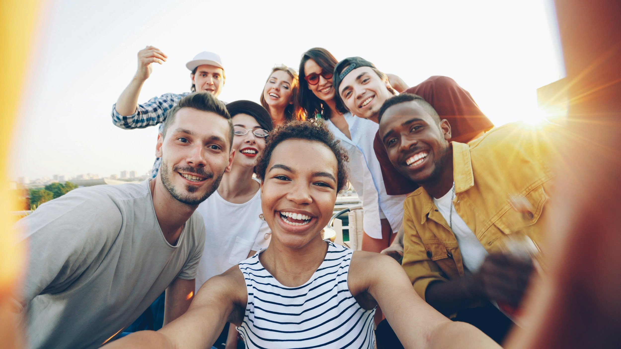 A diverse group of young adult friends smiling and laughing together outdoors as they gather close to take a joyful selfie conveying a sense of community, belonging, and emotional well-being.