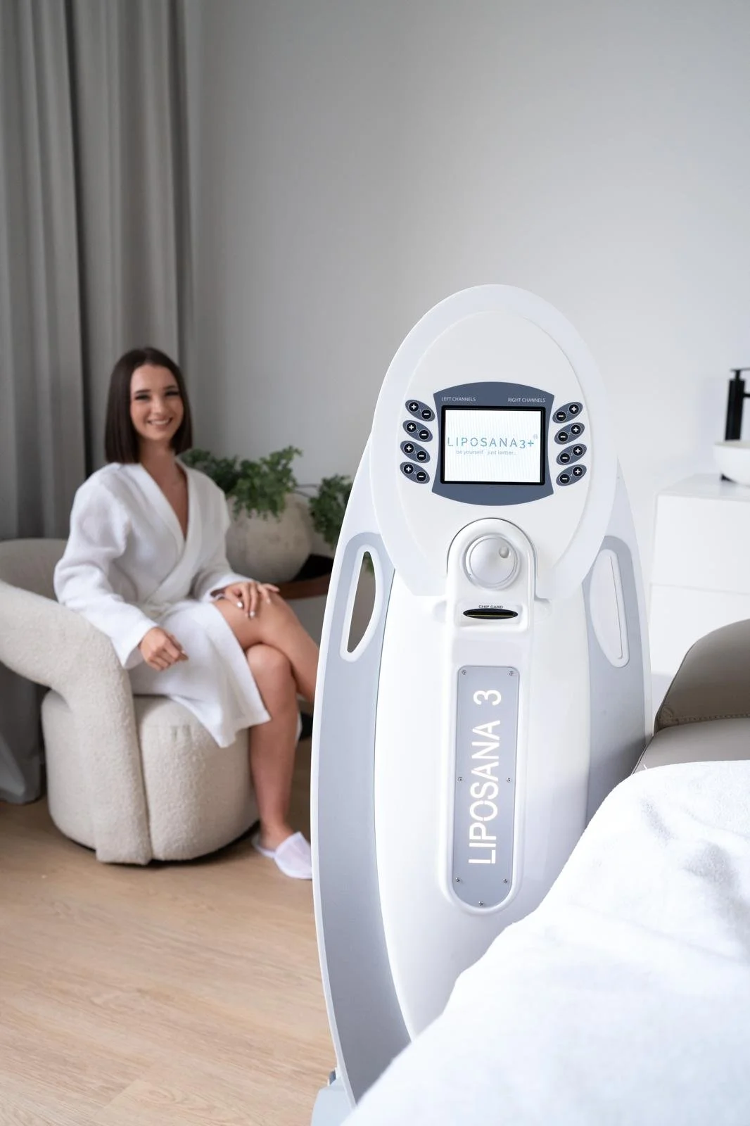 A woman sitting on a beige armchair with a smile, wearing a white robe, beside a modern aesthetic room with potted plants and light-colored curtains, with a large medical or aesthetic device labeled 'LIPOSANA 3' in the foreground.