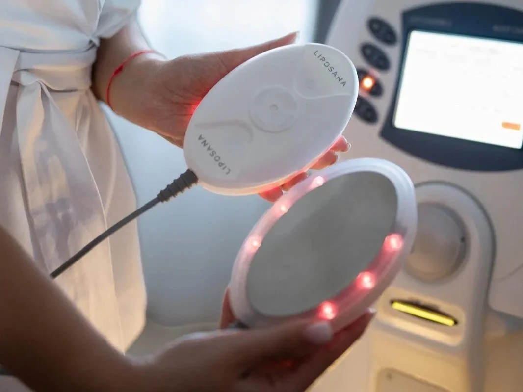 A person holding a portable medical device near a mirror with red LED lights, in a clinical setting with medical equipment and a monitor in the background.