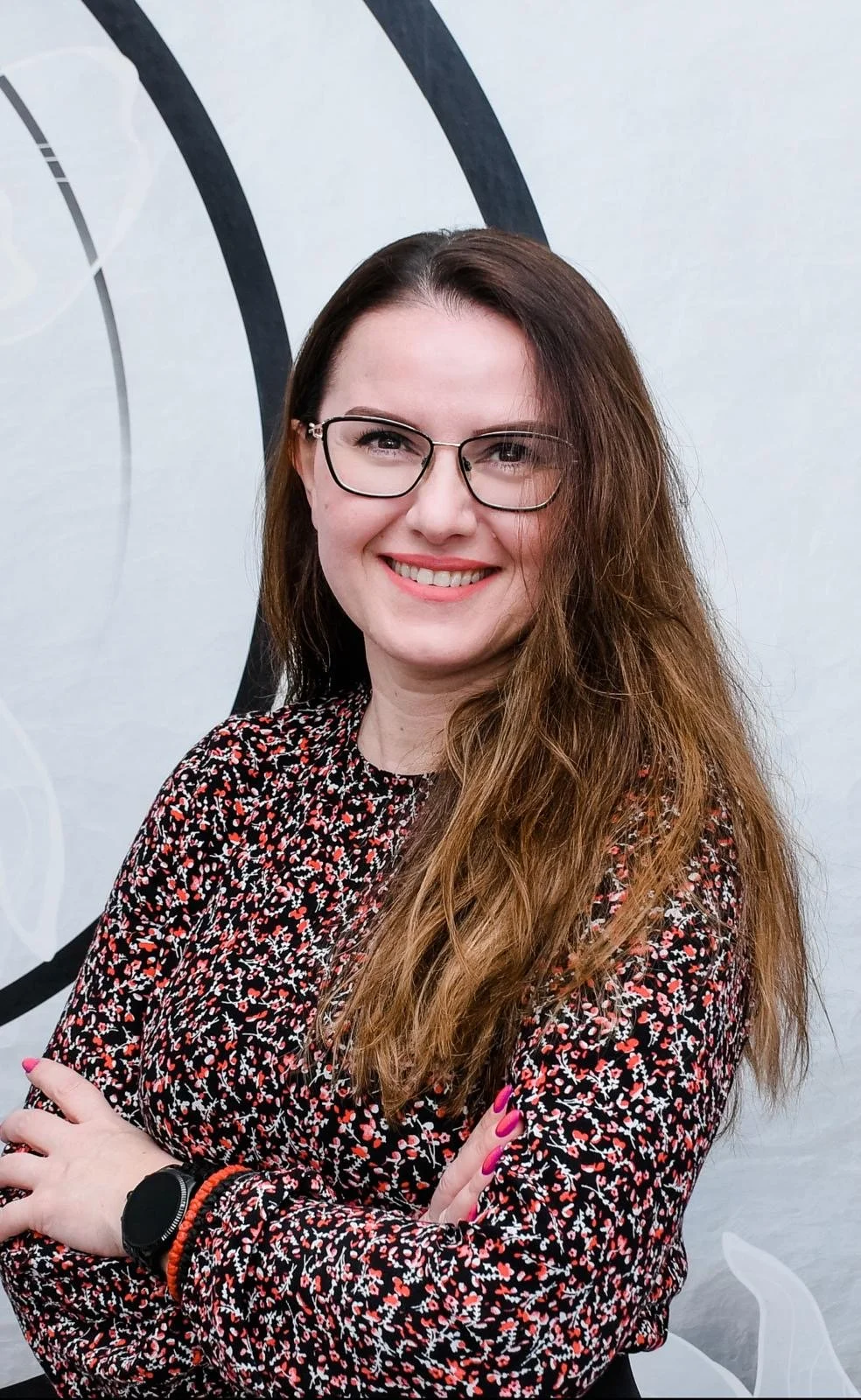 A smiling woman with long, wavy brown hair wearing glasses and a long-sleeved black blouse with a red, white, and pink floral pattern, standing with arms crossed against a light-colored wall with black curved designs.