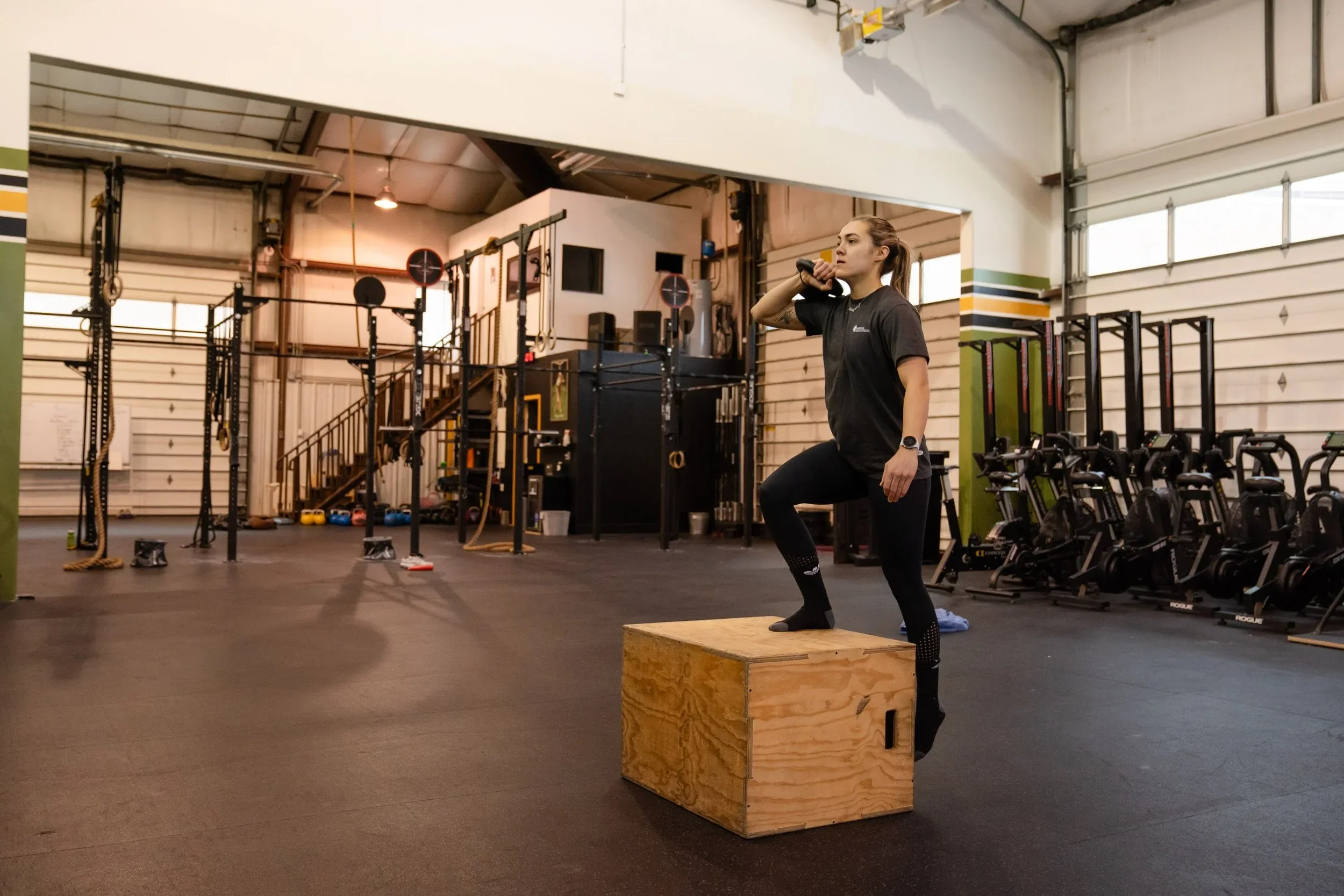 A woman exercises with a kettlebell on her shoulder, standing with one foot on a wooden plyometric box inside a gym.