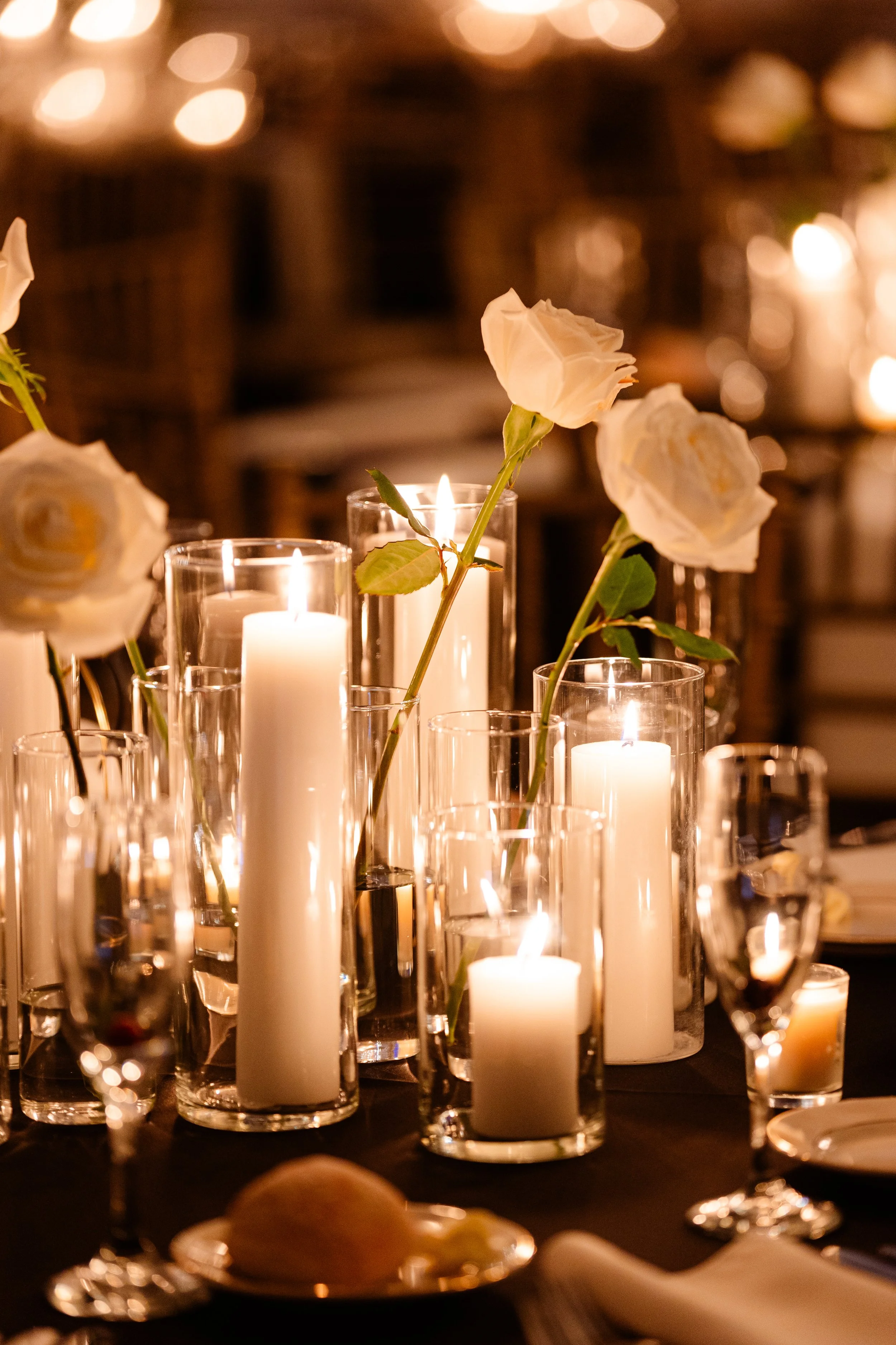 Several white candles and white roses in glass vases arranged on a table with dim lighting.