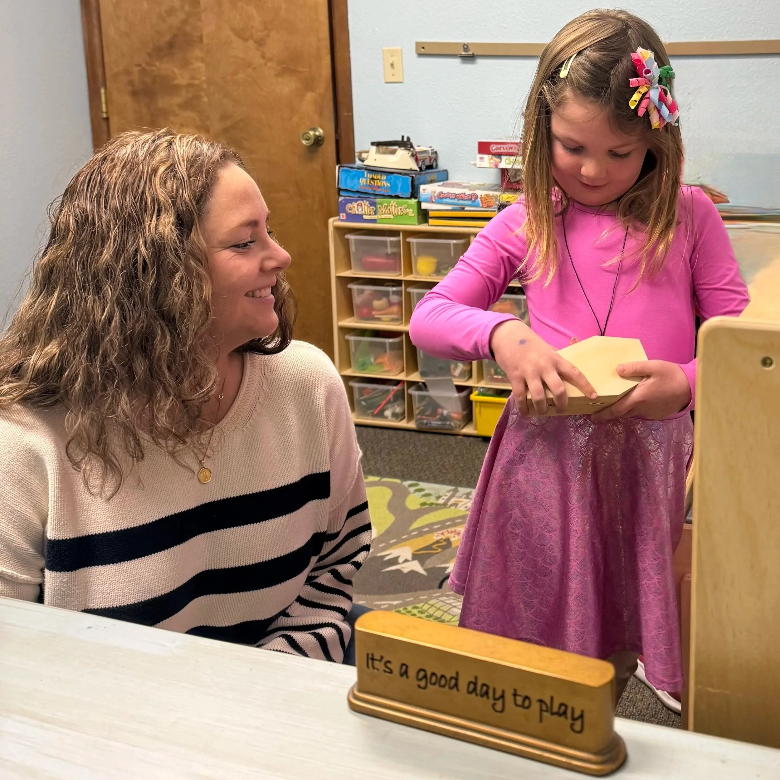 A woman with curly hair wearing a striped sweater is smiling at a young girl with a colorful bow in her hair, who is holding a wooden block in a classroom setting. There is a sign on the table that says, 'It's a good day to play.'