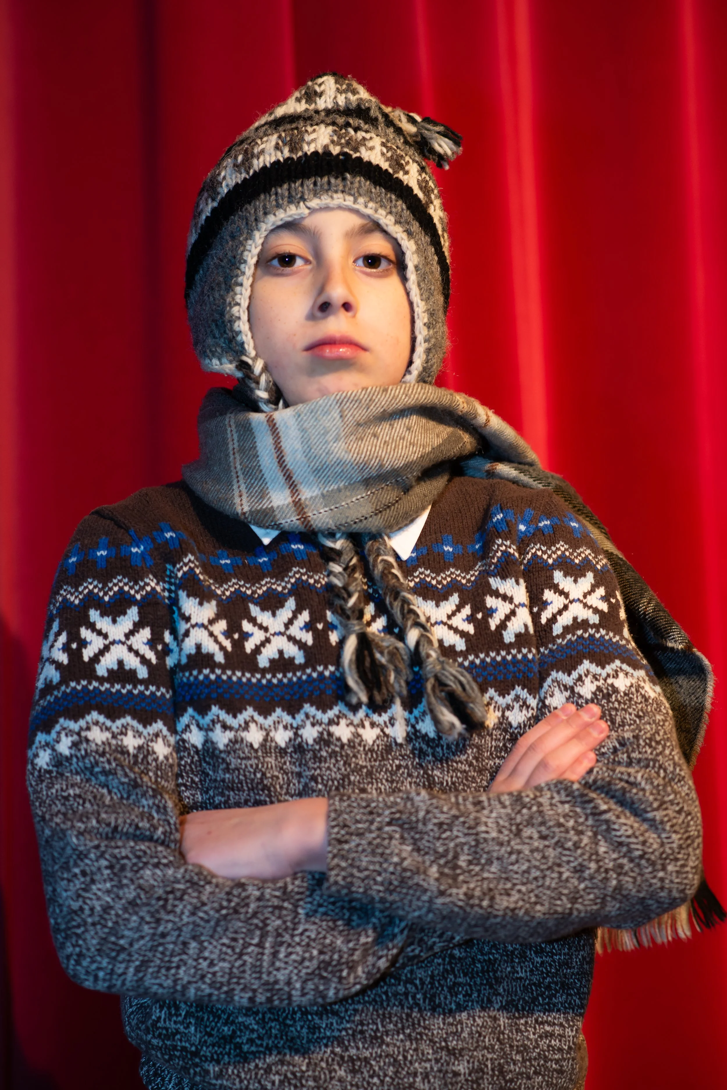 A young boy wearing a patterned knit hat, a beige scarf, and a brown sweater with snowflake designs, standing with crossed arms in front of a red curtain.