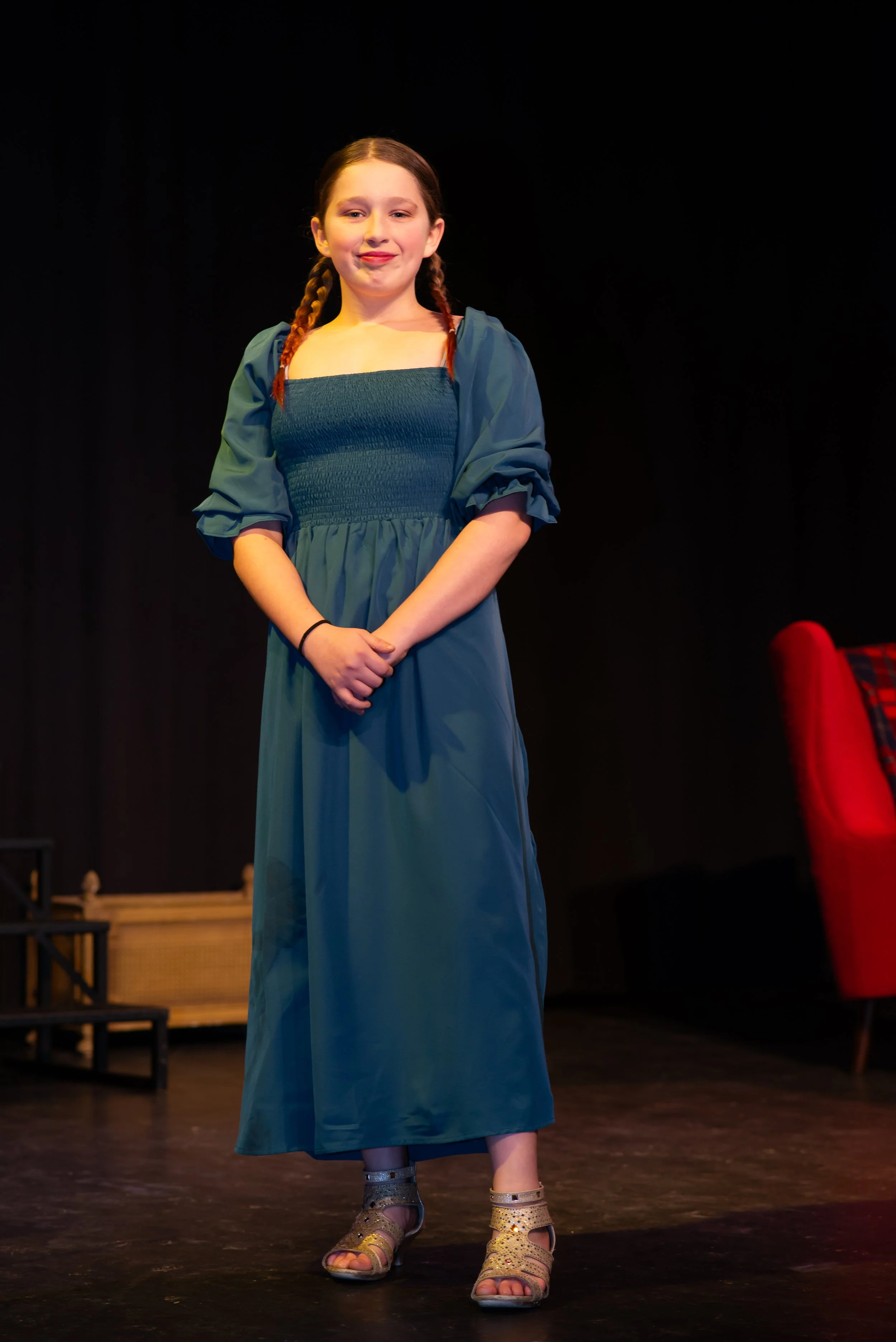 A young girl with braided hair standing on a stage, wearing a long blue dress and gold sandals.