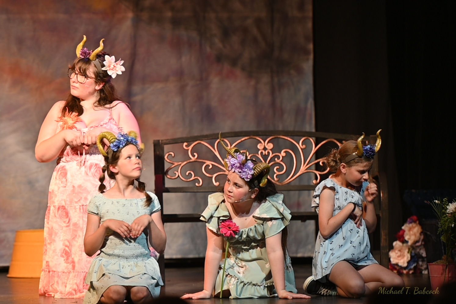 Scene from a theatrical play featuring five young girls dressed as mythical creatures with horns, floral accessories, and costumes, performing on stage with a decorative background.