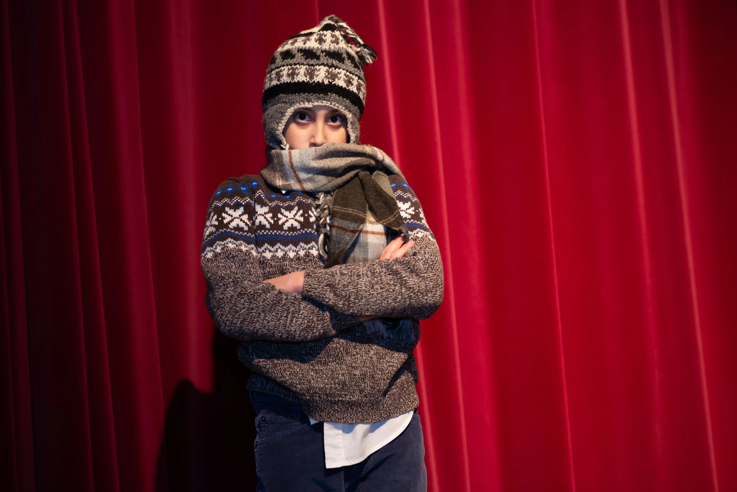A young boy wearing a winter hat, scarf, and sweater standing in front of red velvet curtains.