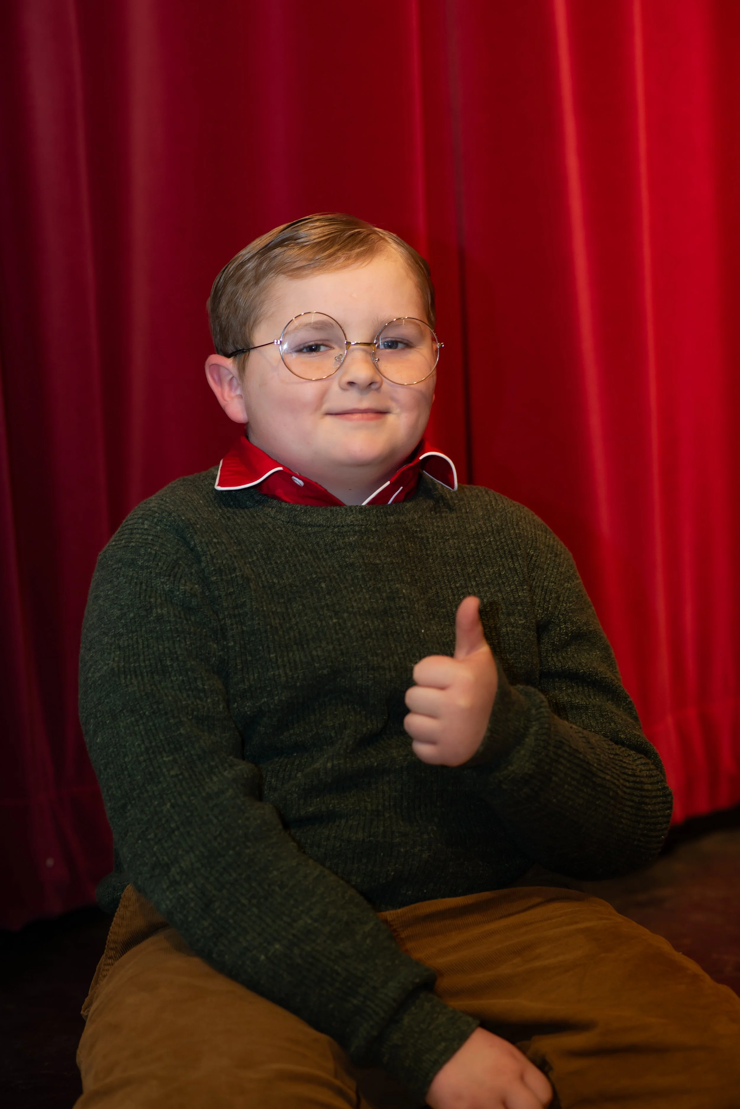 A young boy with light brown hair, wearing glasses, a red shirt, and a green sweater, sitting against a red curtain background, giving a thumbs-up.