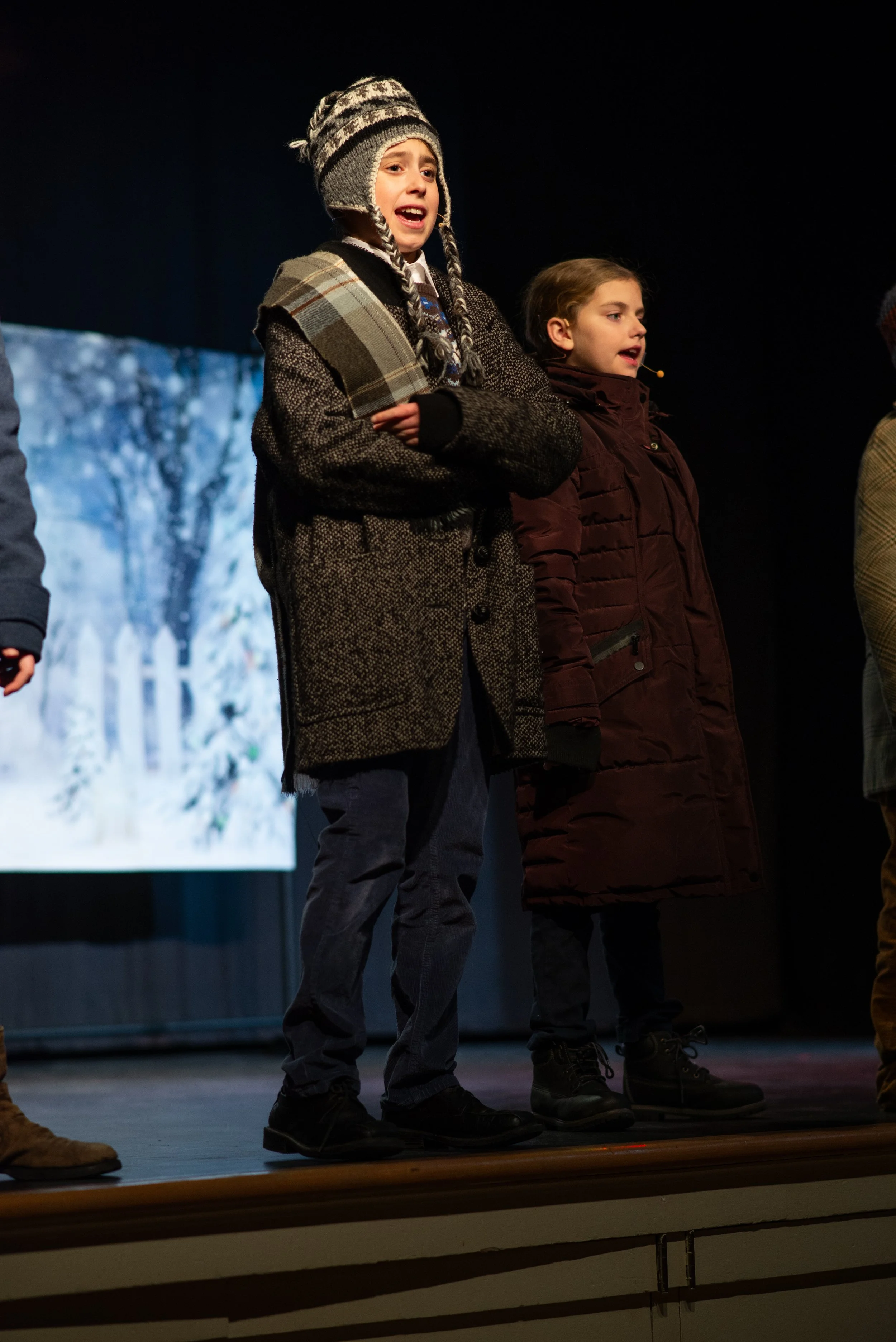 Two children standing on a stage, possibly performing or speaking, with a snowy winter scene projected on a screen behind them. The child on the left is wearing a plaid scarf and a knit hat, and the child on the right is wearing a brown coat.