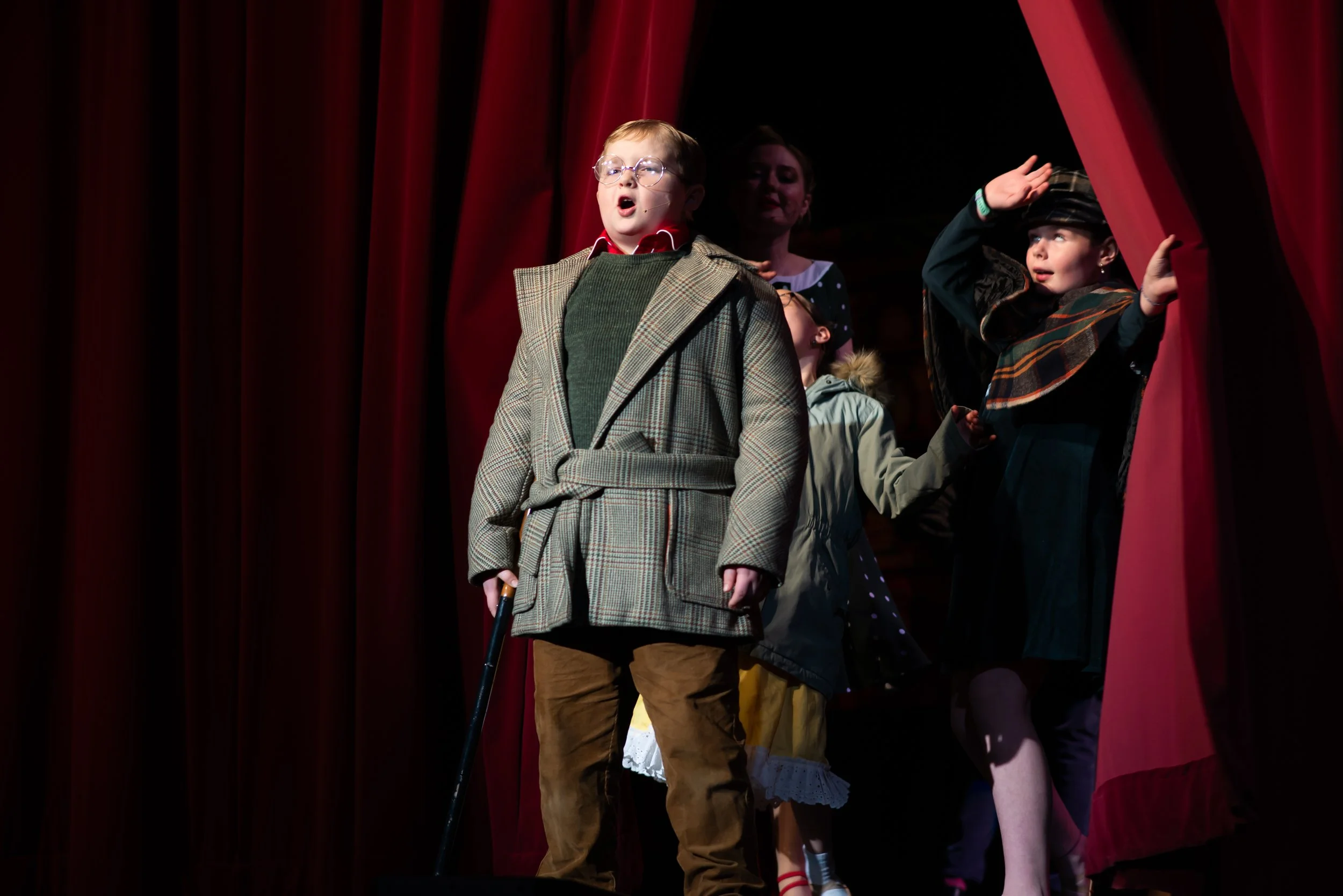Children dressed in costumes on stage behind red curtains, performing or preparing to perform.