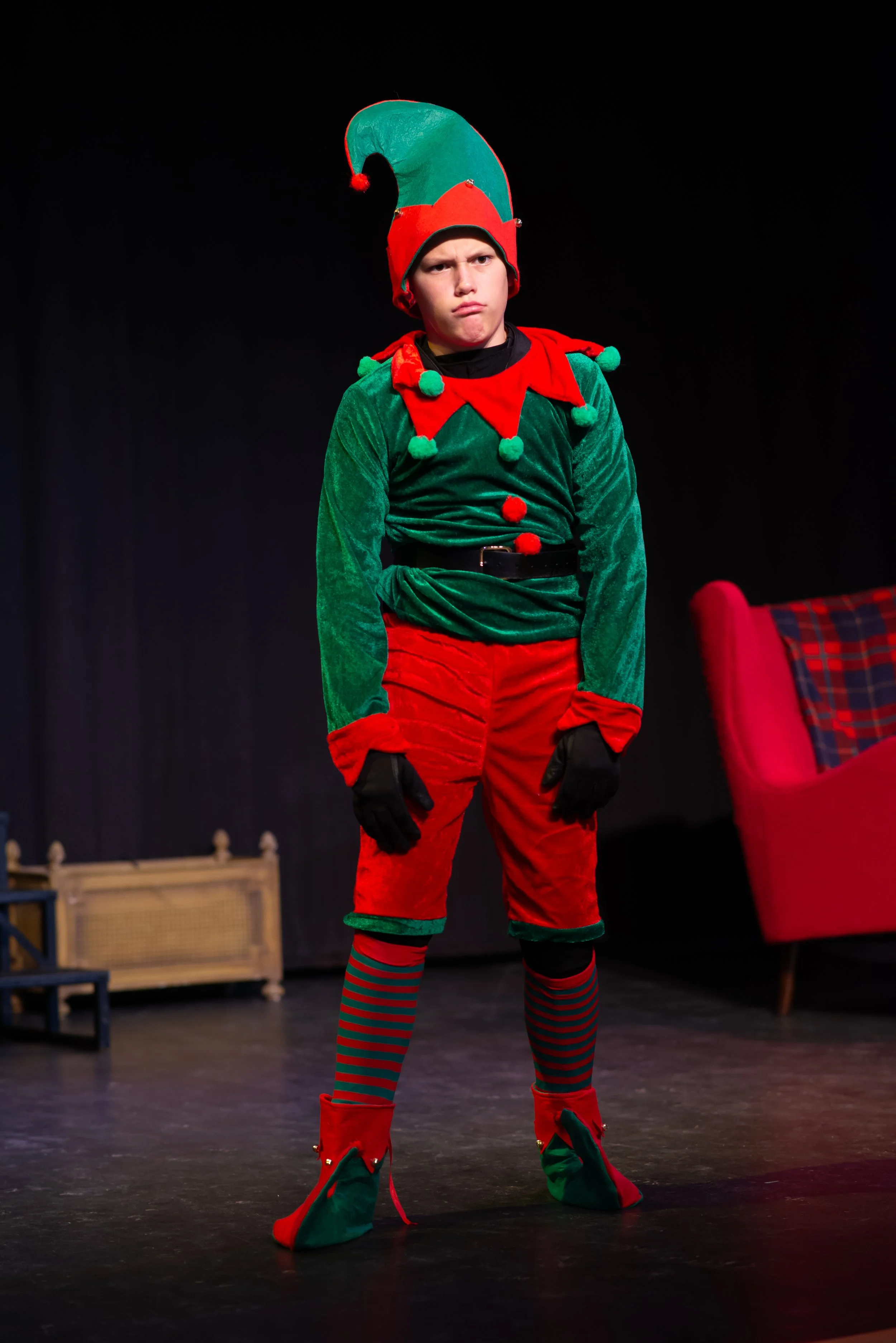 A young person dressed as an elf with a red and green hat with a pom-pom, a green velvet shirt with red accents, red pants, and elf shoes, standing on a stage.