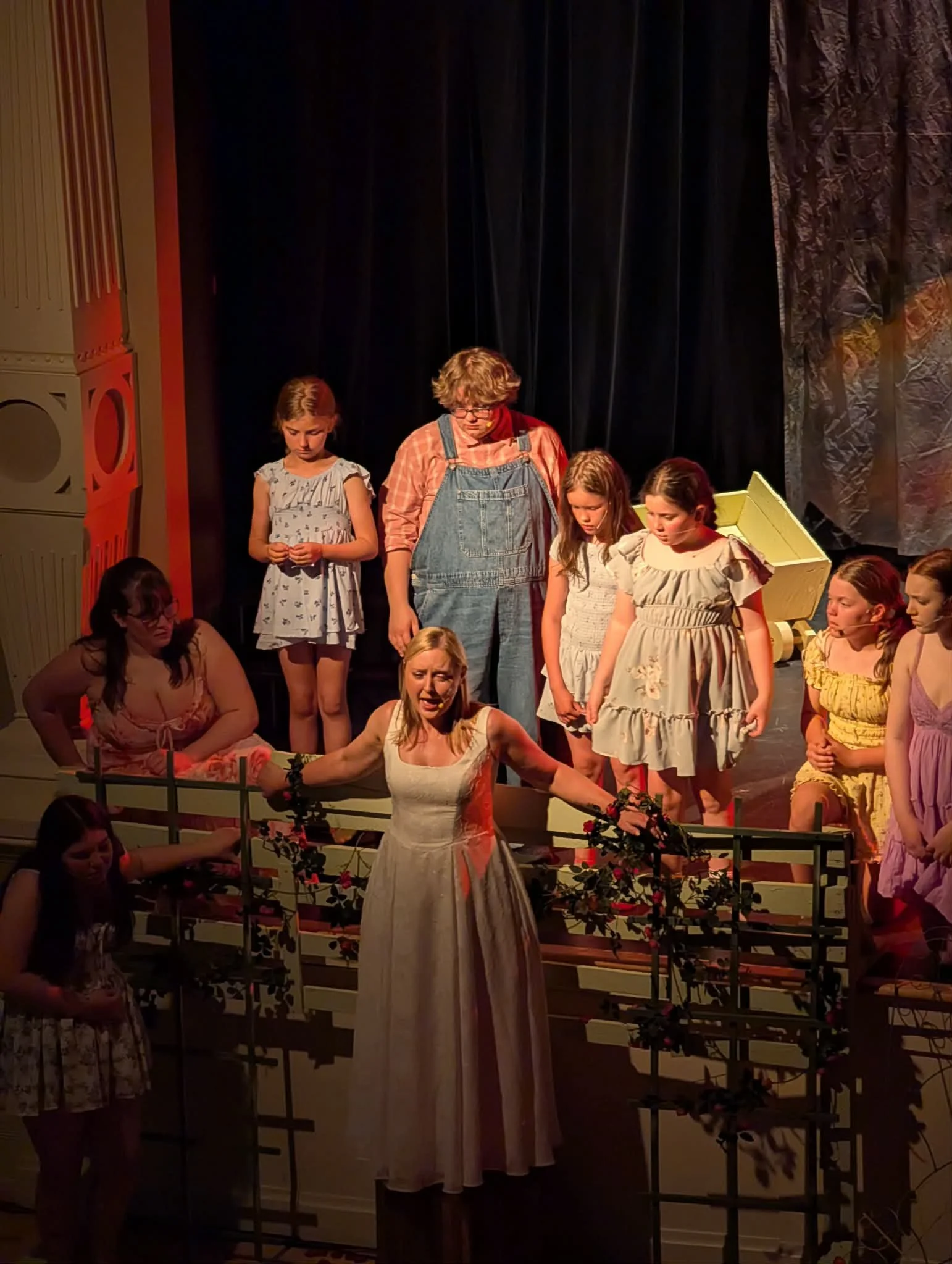 Children and an adult are standing on a stage during a theatrical performance, with black curtains and stage props around them.
