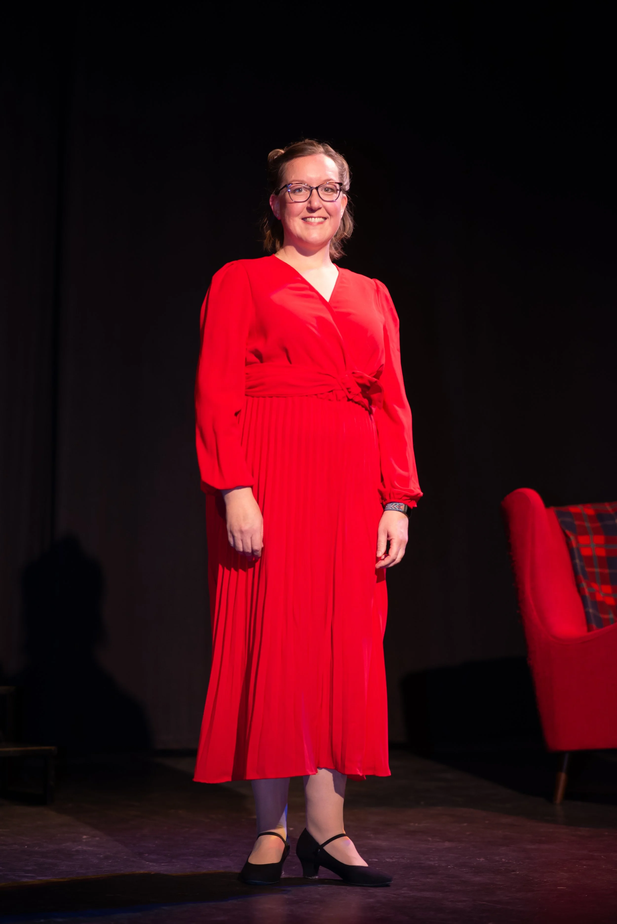A woman dressed in a bright red, long-sleeved gown with pleats, standing on stage with a dark background. She is wearing glasses, smiling, and has her hair styled back. She is also wearing black shoes with ankle straps.