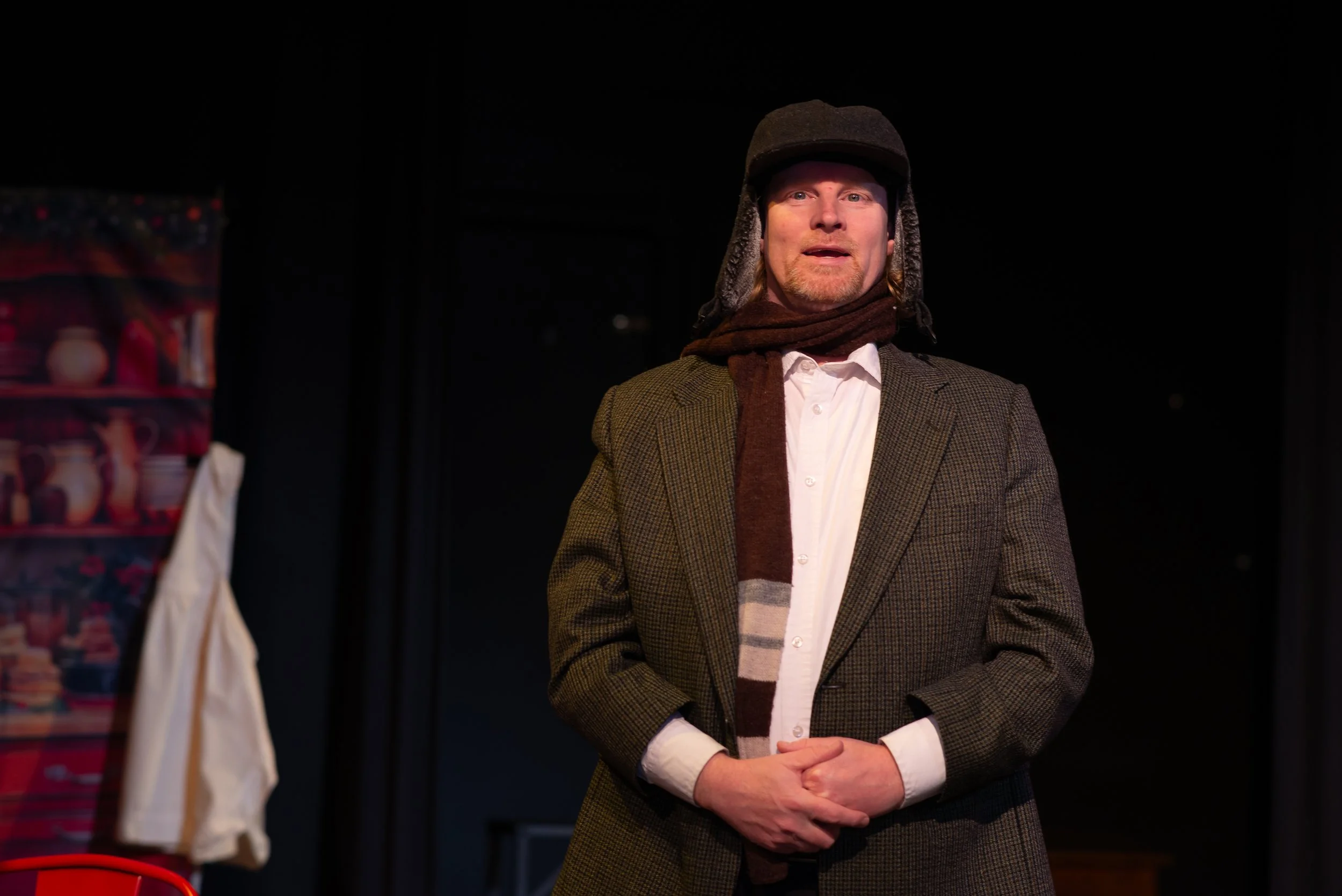 Man wearing a plaid blazer, white shirt, and a winter hat with ear flaps, standing with hands clasped in front, in a dark setting, possibly on stage.