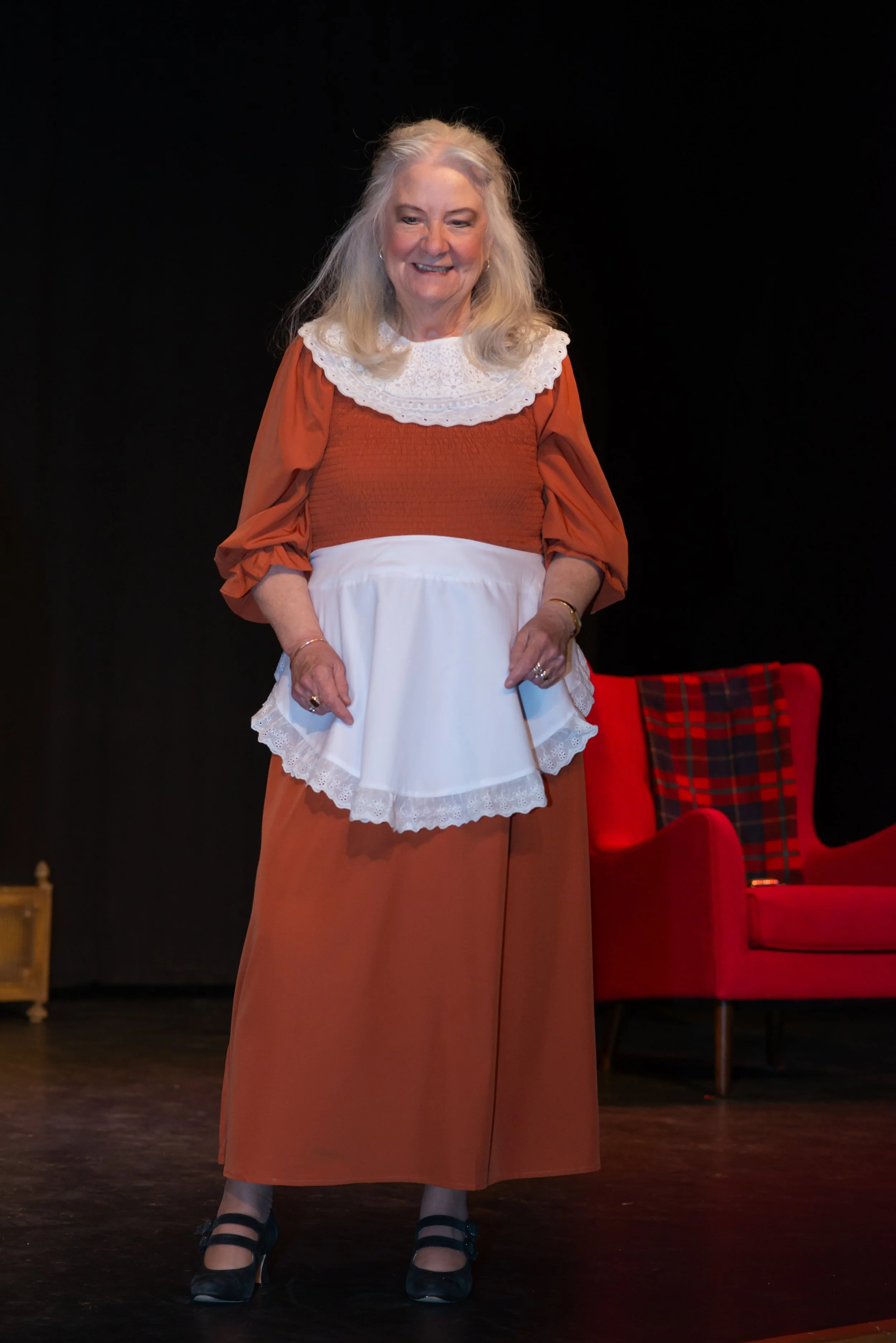 A woman with long gray hair wearing a vintage-inspired brown dress with puffed sleeves, a white lace apron, and a white lace collar, standing on a stage with a black background and red chairs.