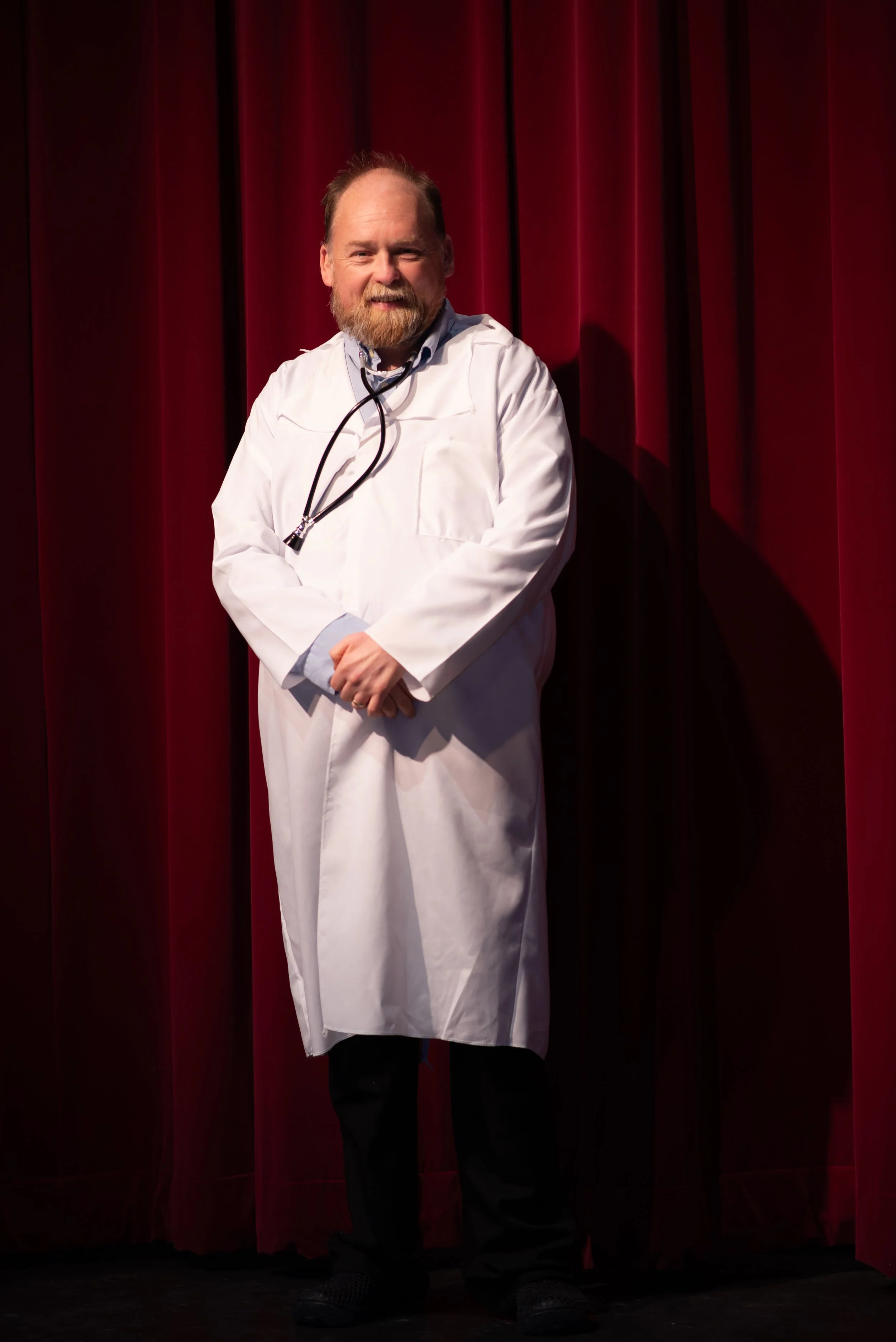 A man with a beard and mustache, dressed in a white coat and black shoes, standing in front of red stage curtains, smiling and looking at the camera.