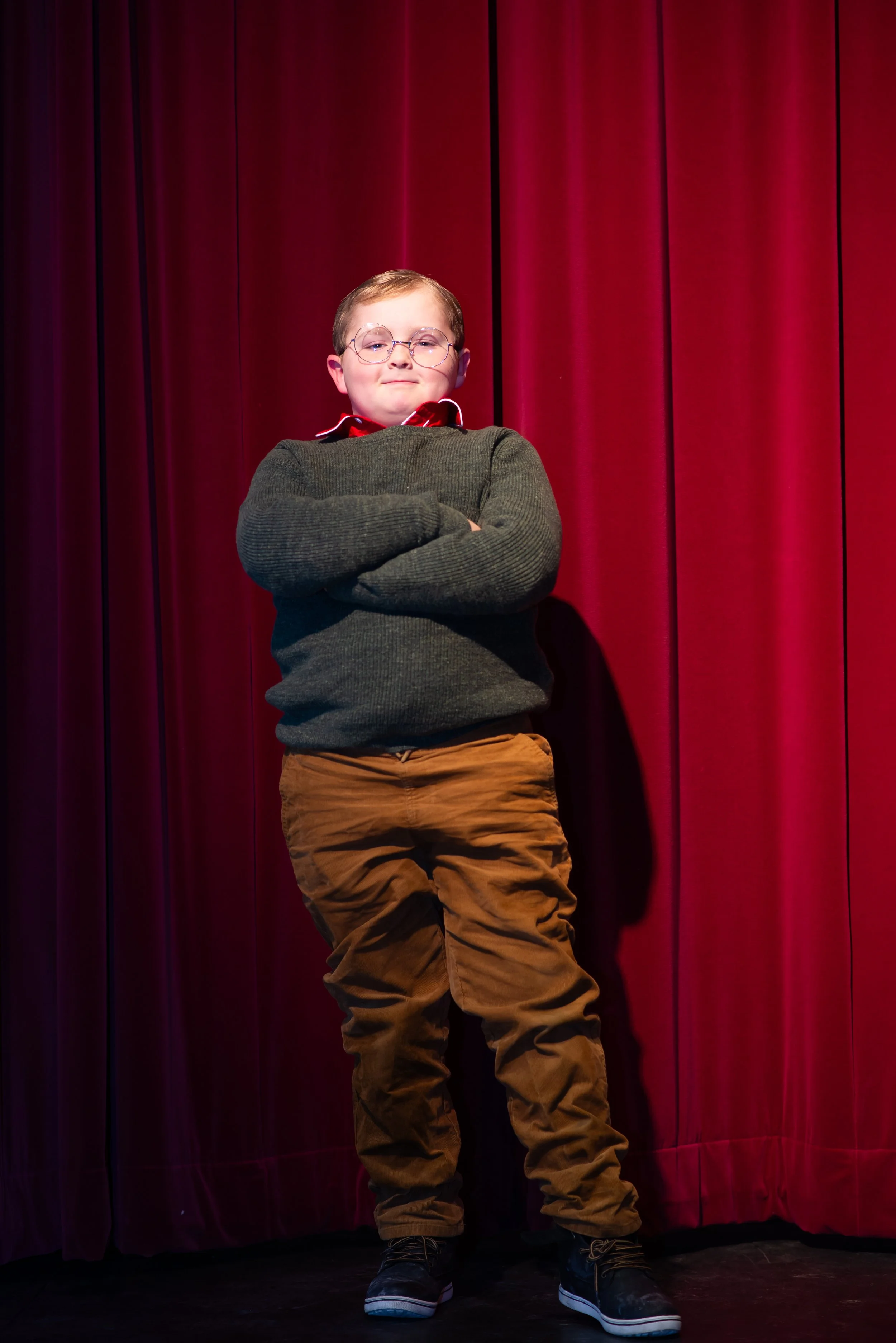 A young boy with glasses, wearing a dark sweater, orange pants, and black shoes, standing in front of a red curtain with arms crossed.
